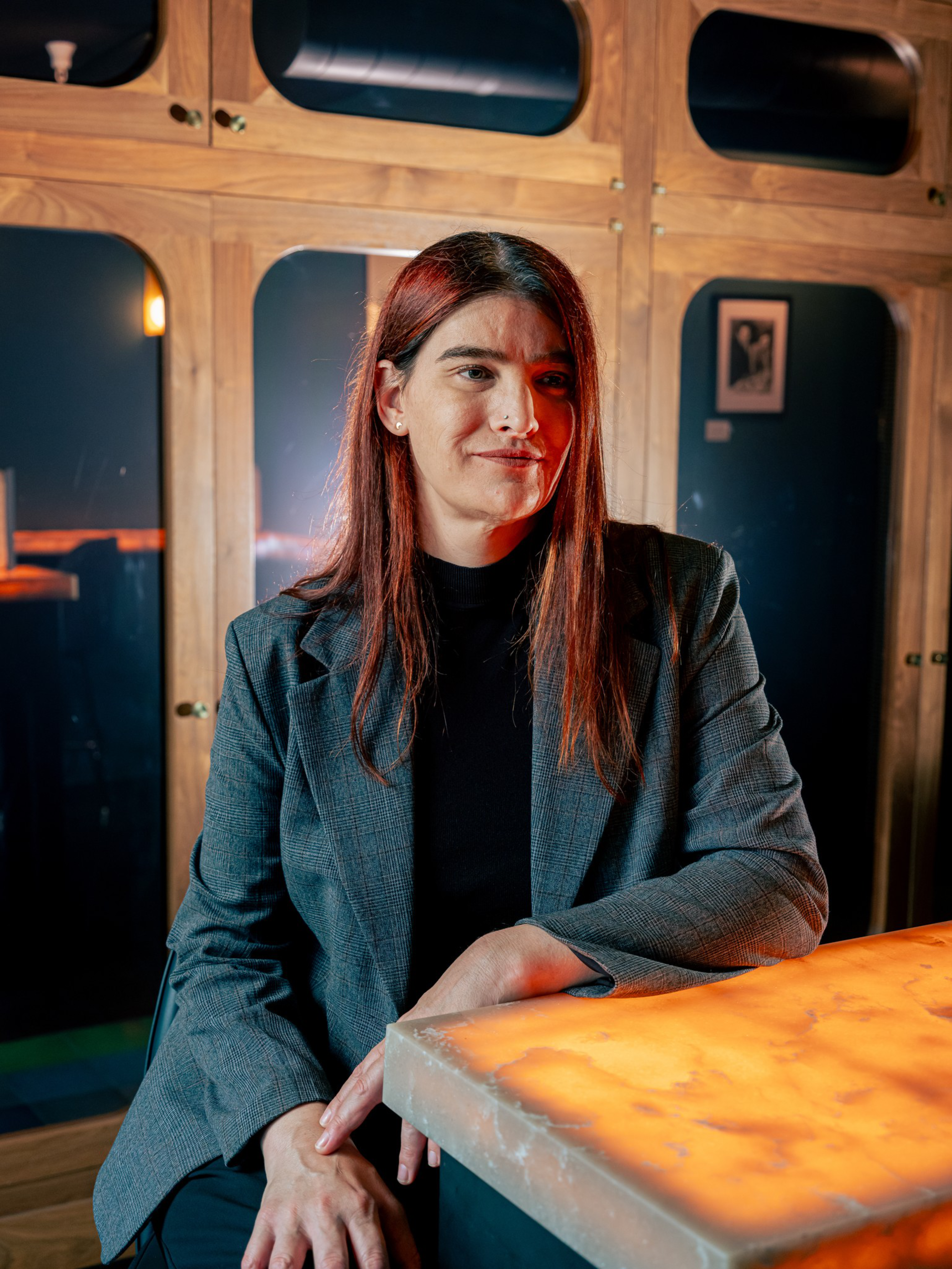 A woman with long brown hair wears a gray blazer and black shirt, sitting at a warm-lit marble table against wooden cabinets and dark blue walls.