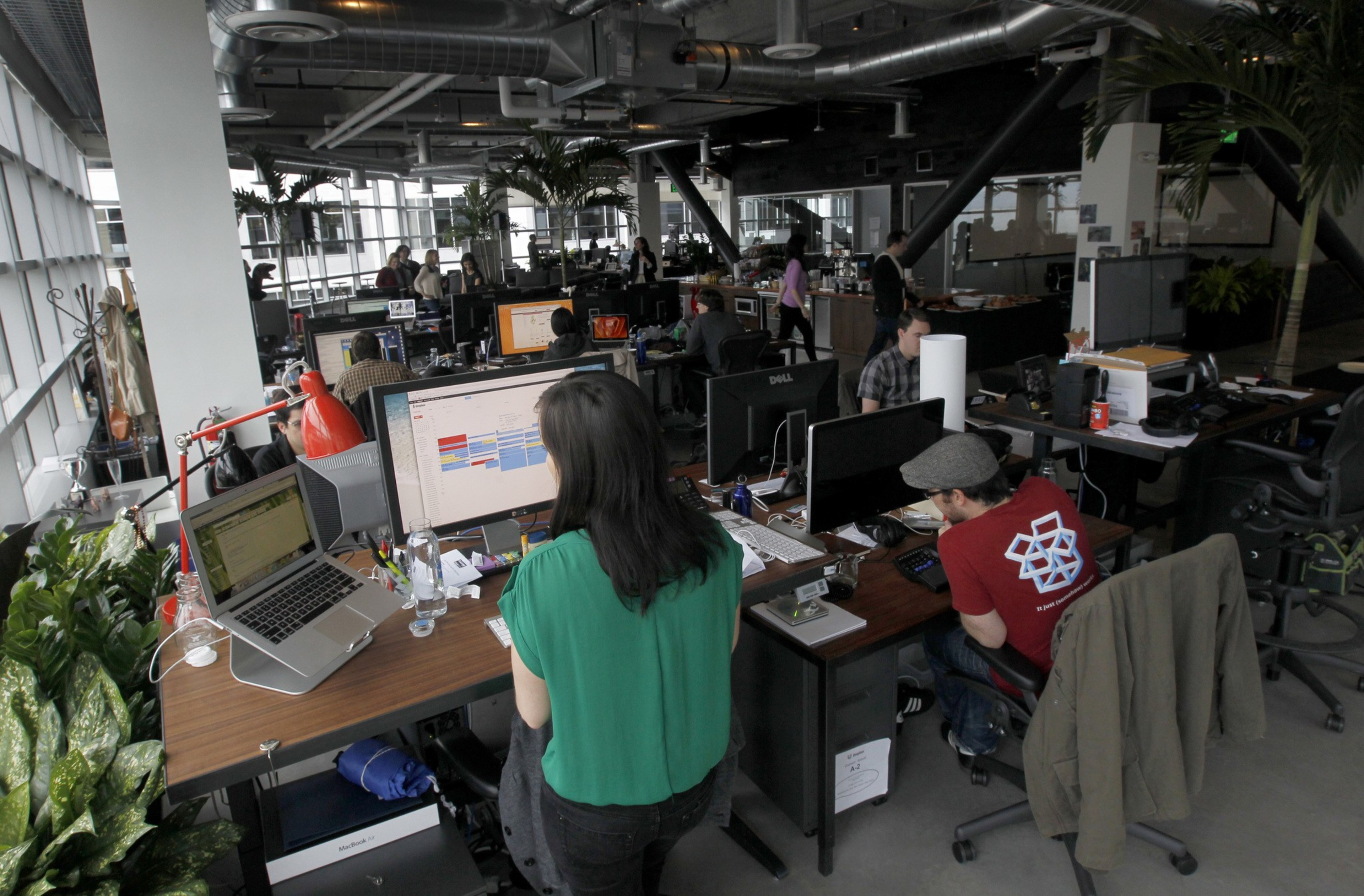 An office with people working at cluttered desks, computers everywhere, and large windows.