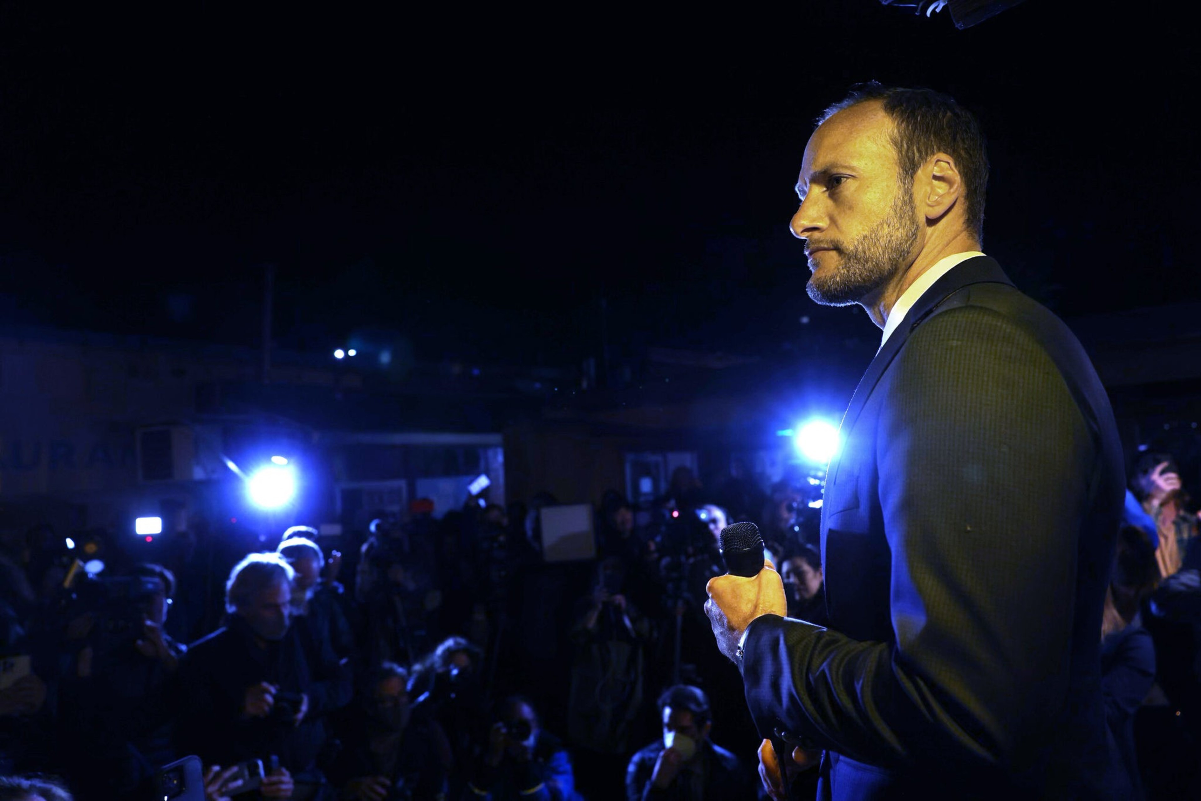 Chesa Boudin stands before a crowd, facing bright lights, holding a microphone.