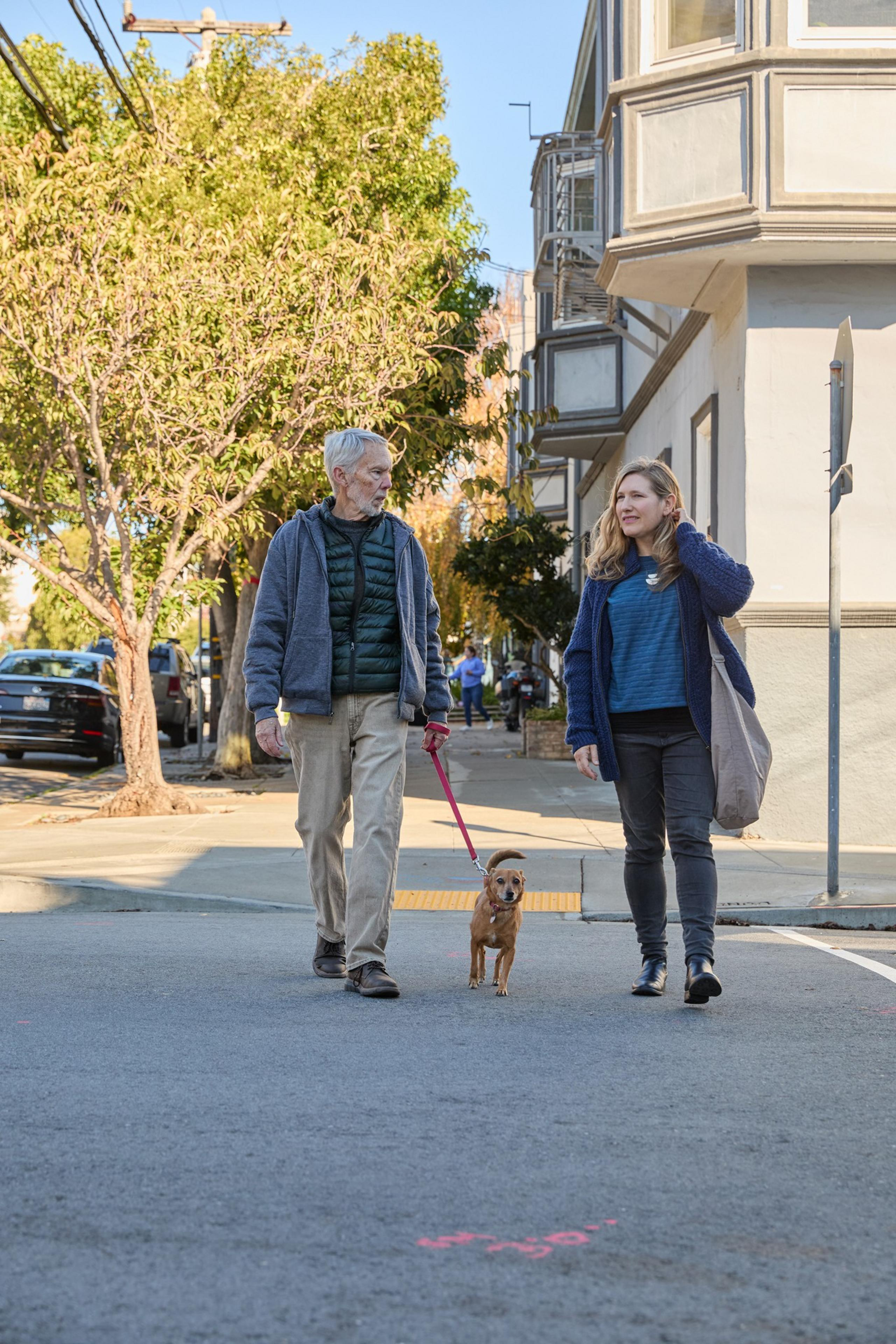 An older man and a younger woman walk a small brown dog down a tree-lined street. They wear casual clothes, and the scene is set in a residential area.