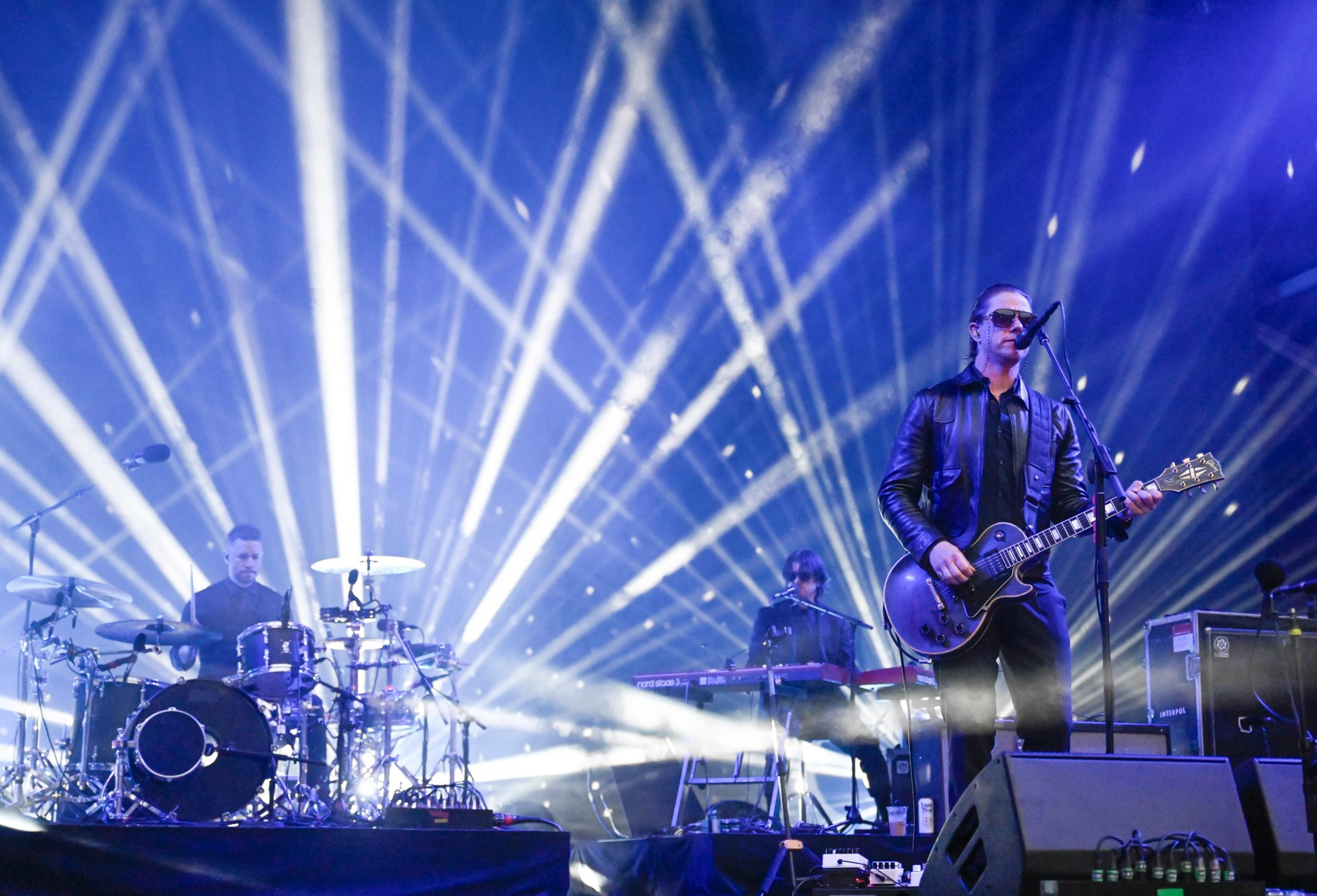 A band performs on stage with a guitarist and singer in front, a drummer on the left, and a keyboardist in the center, all under bright white stage lights.