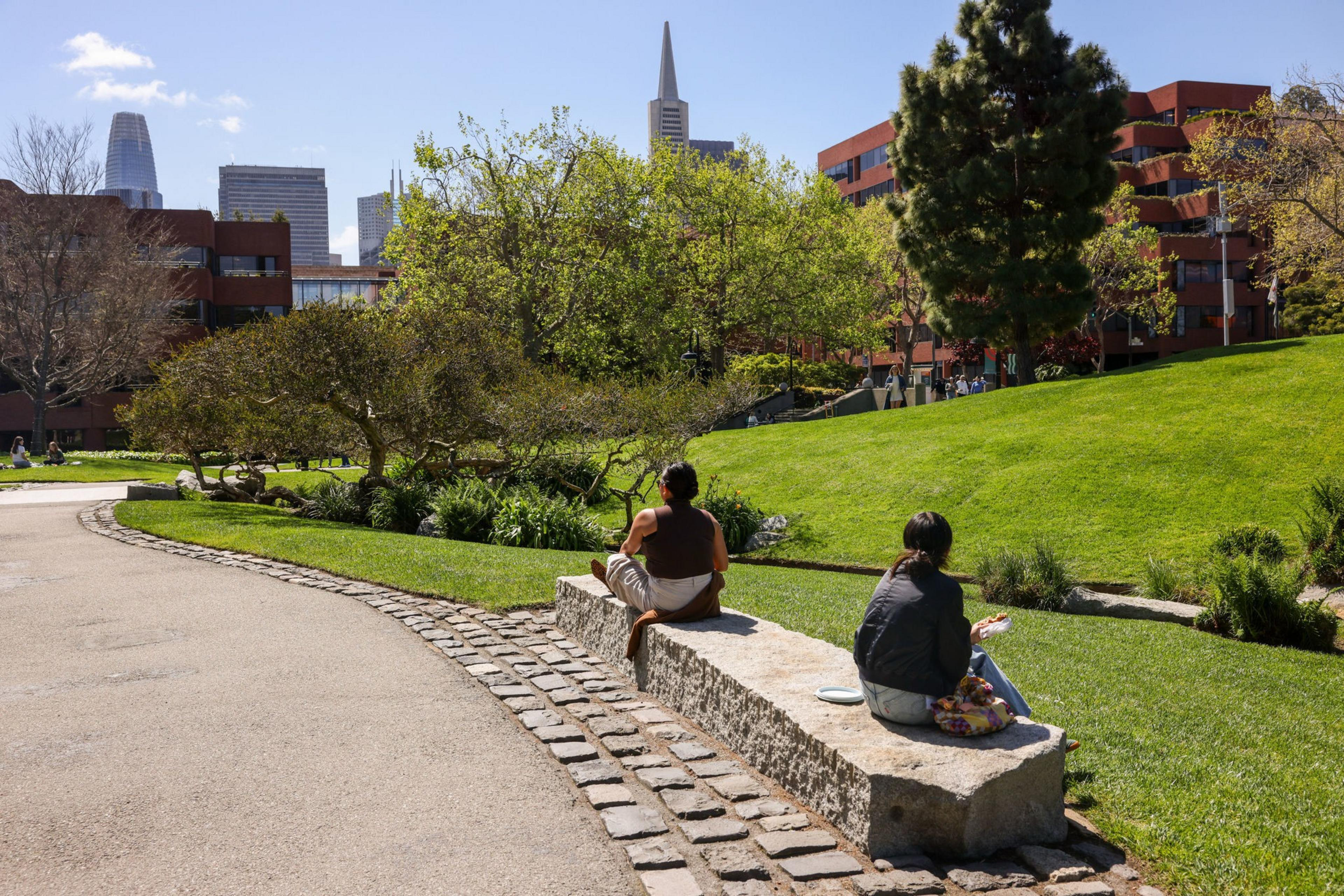 Two people sit on a stone bench in a park with green grass and trees. Modern buildings are visible in the background under a clear blue sky.