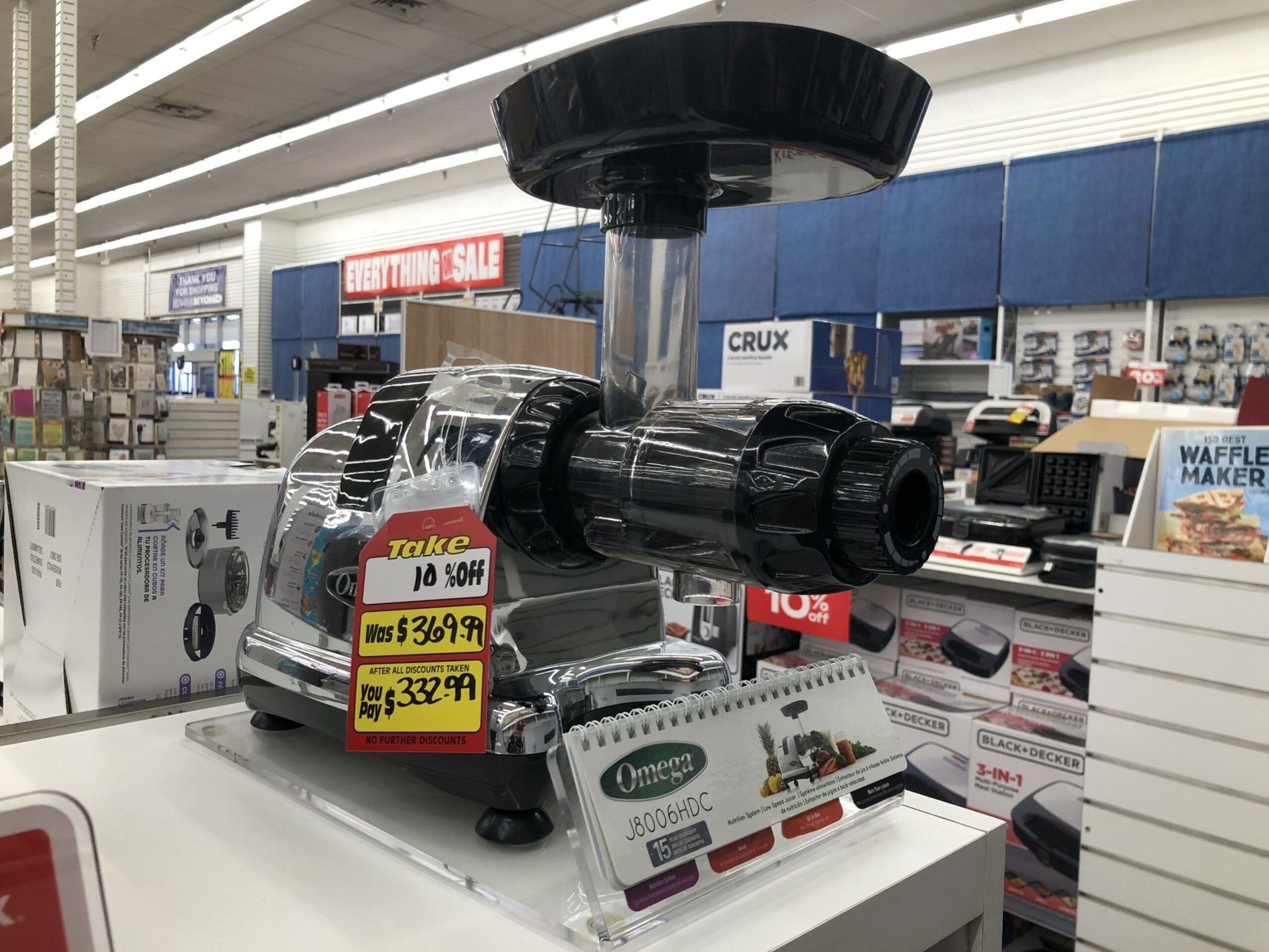 A shiny black and silver Omega juicer sits on a white counter with a 10% off sale tag showing a price reduction to $332.99. Various kitchen appliances are visible in the background.