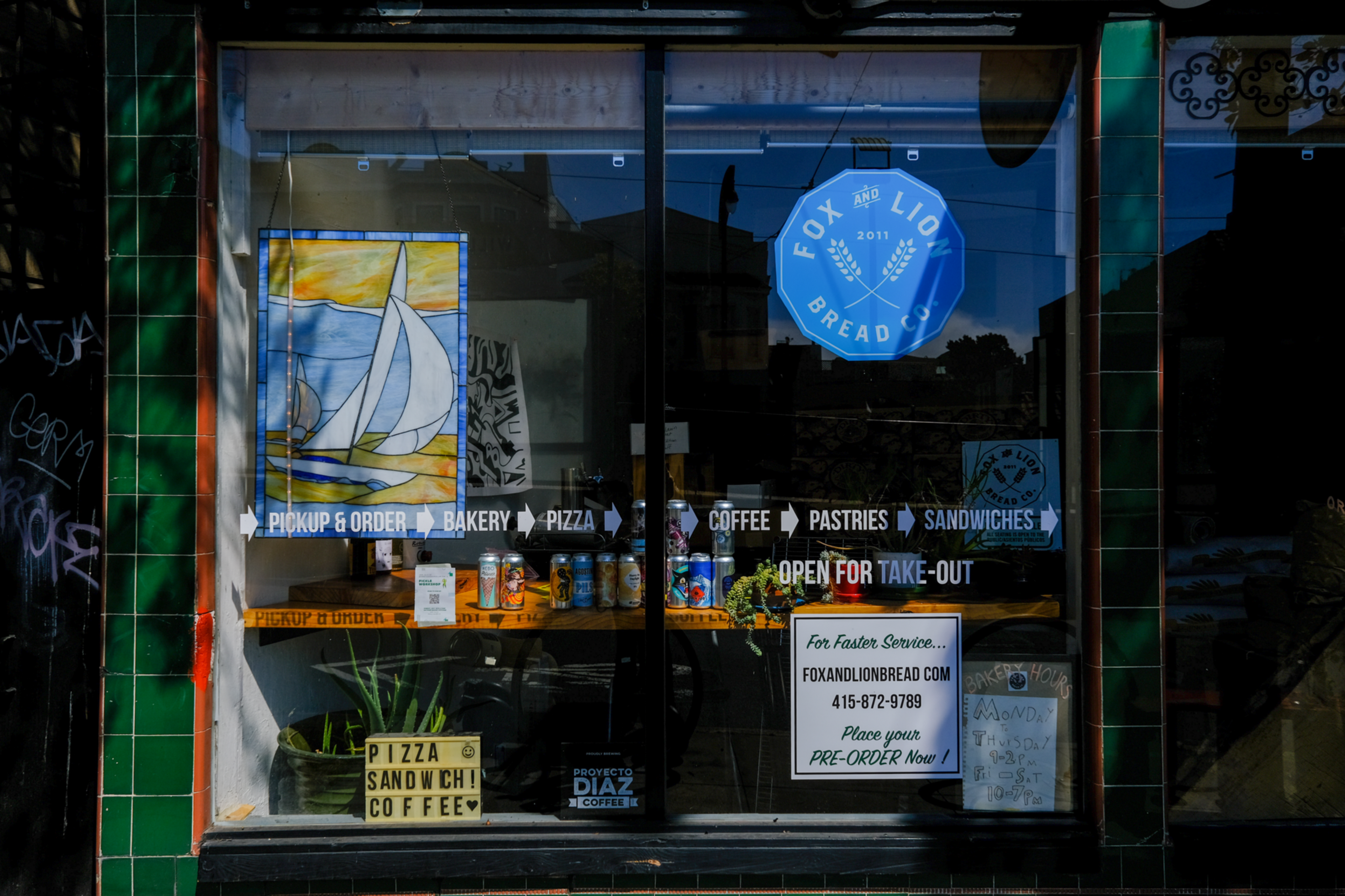 A bakery and coffee shop window displays a sailboat painting, drink cans, and signs for pizza, sandwiches, coffee, pastries, and take-out orders.