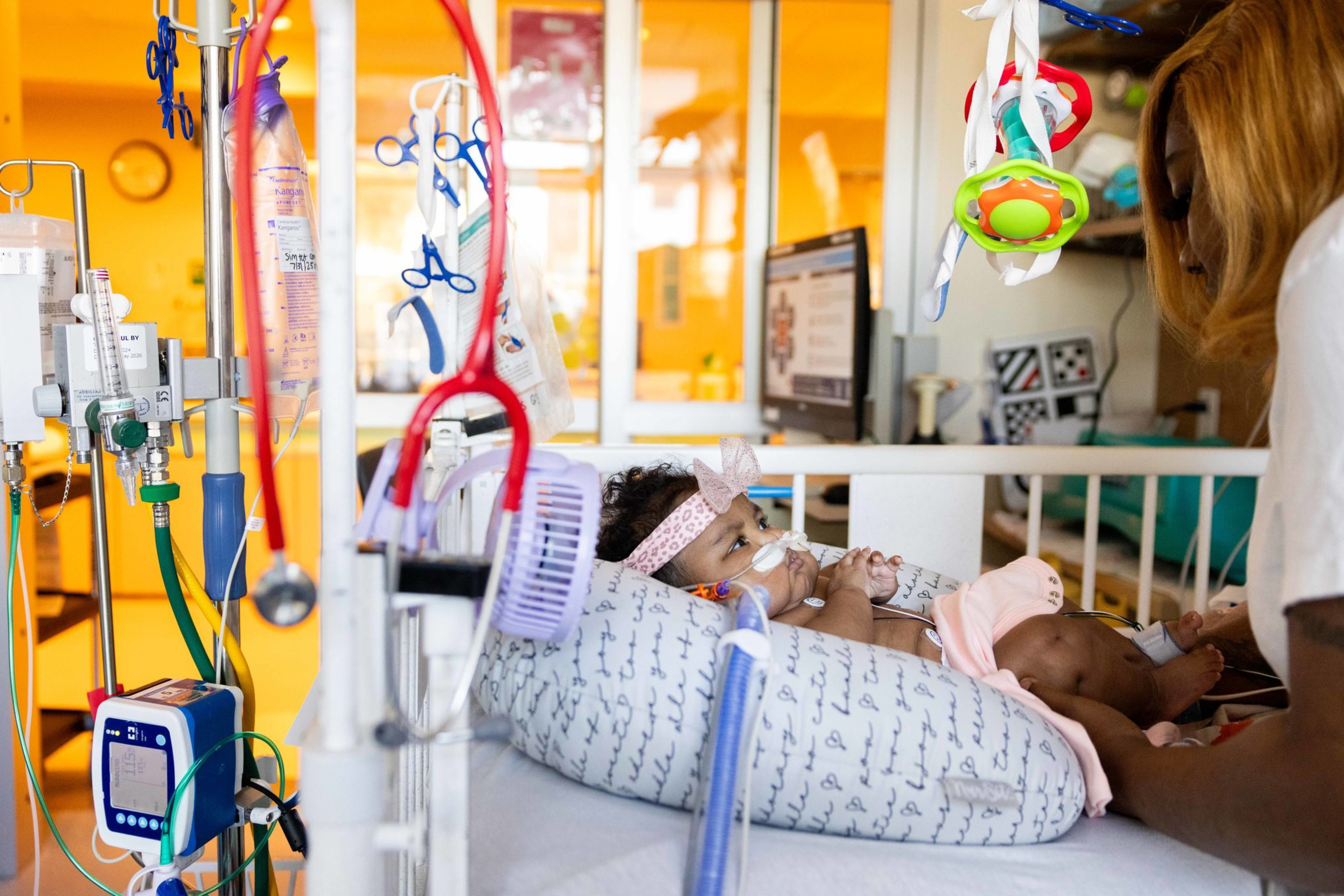 A baby lies in a hospital crib with medical tubes, wearing a breathing mask and headband, while a woman attentively holds the baby's hands.