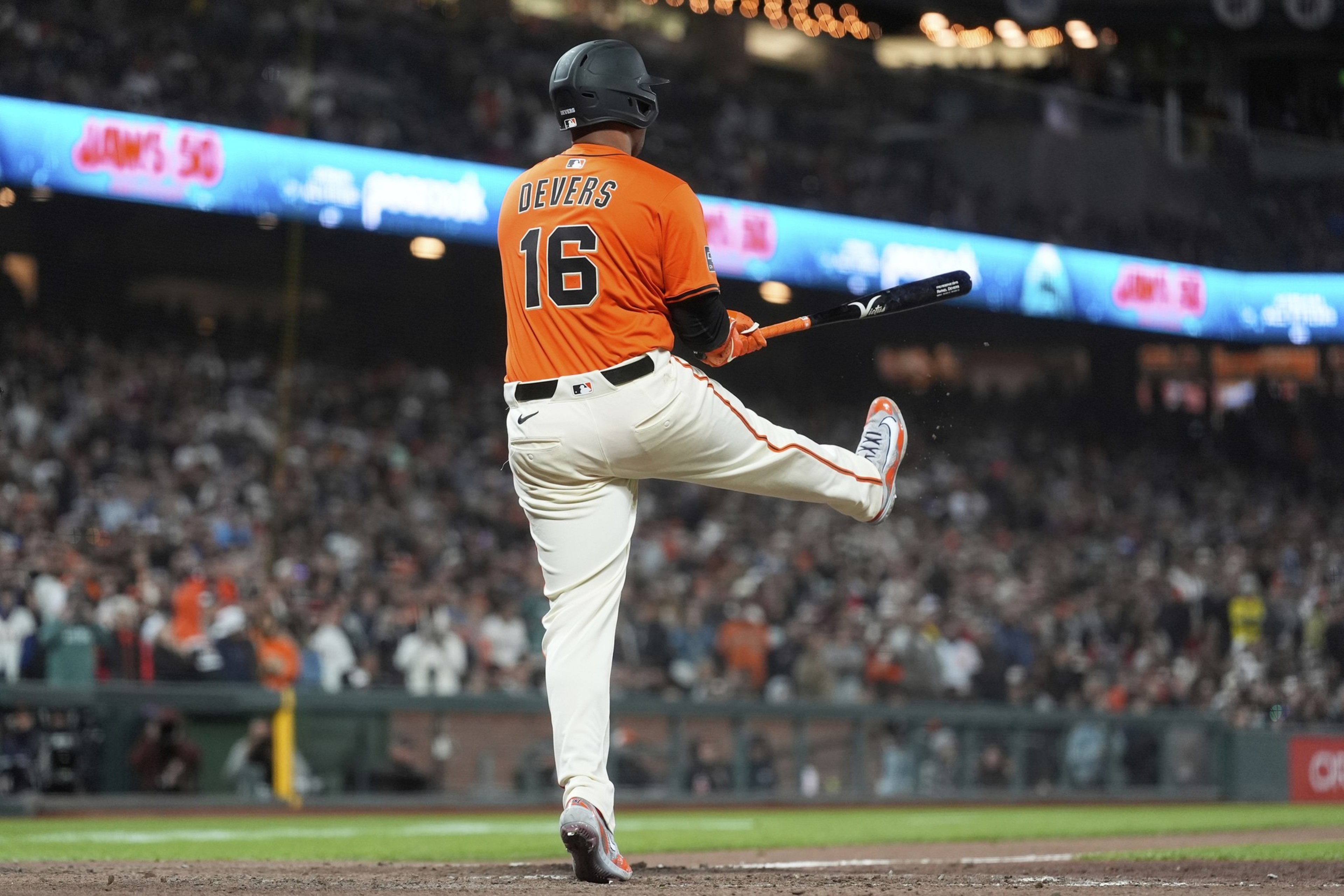 A baseball player in an orange jersey, number 16, raises his leg while holding a bat at a crowded stadium, with bright lights and signs in the background.