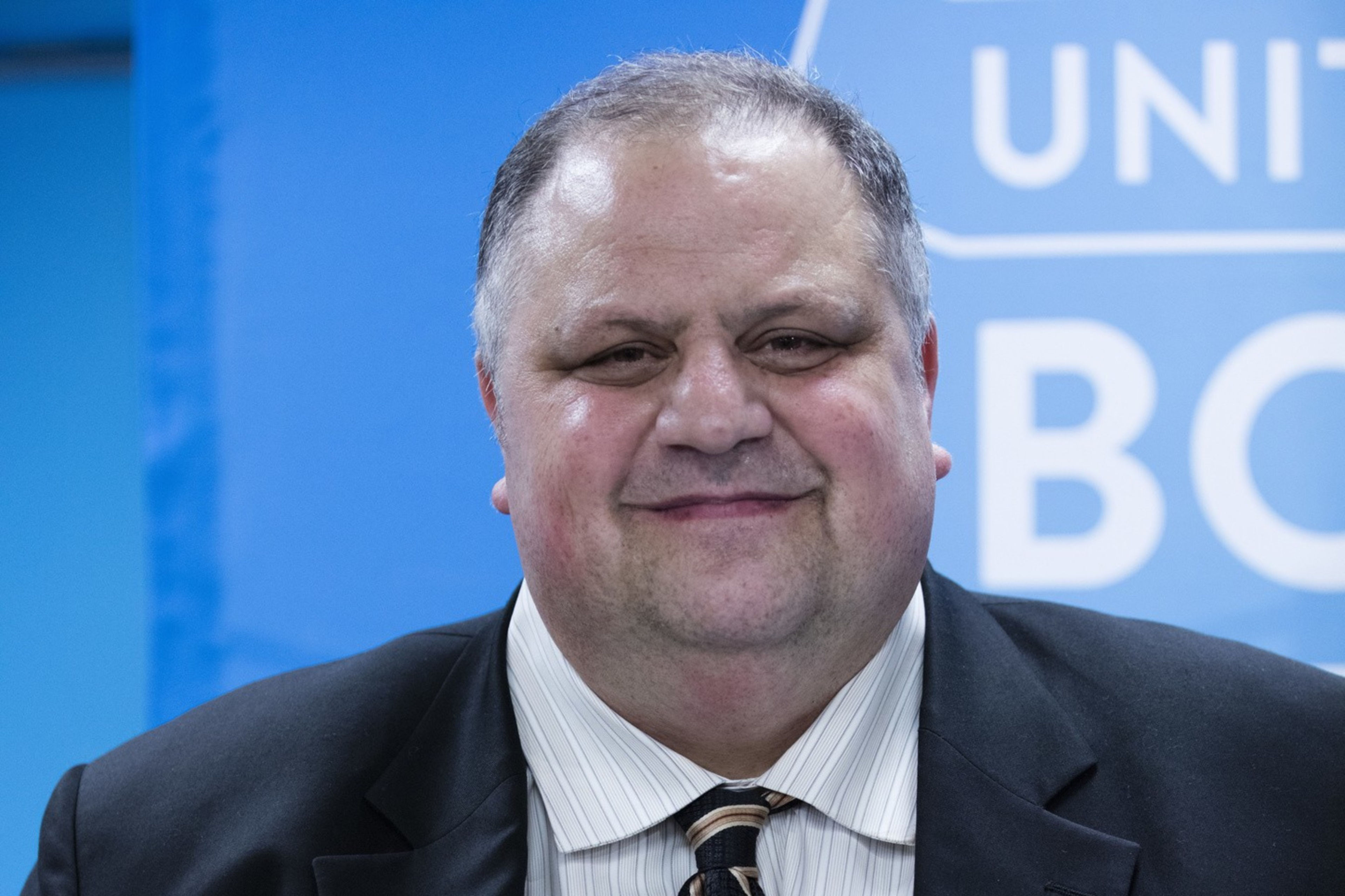 A man in a suit holds the book "NeuroTribes" by Steve Silberman, standing against a blue background with partial words visible.
