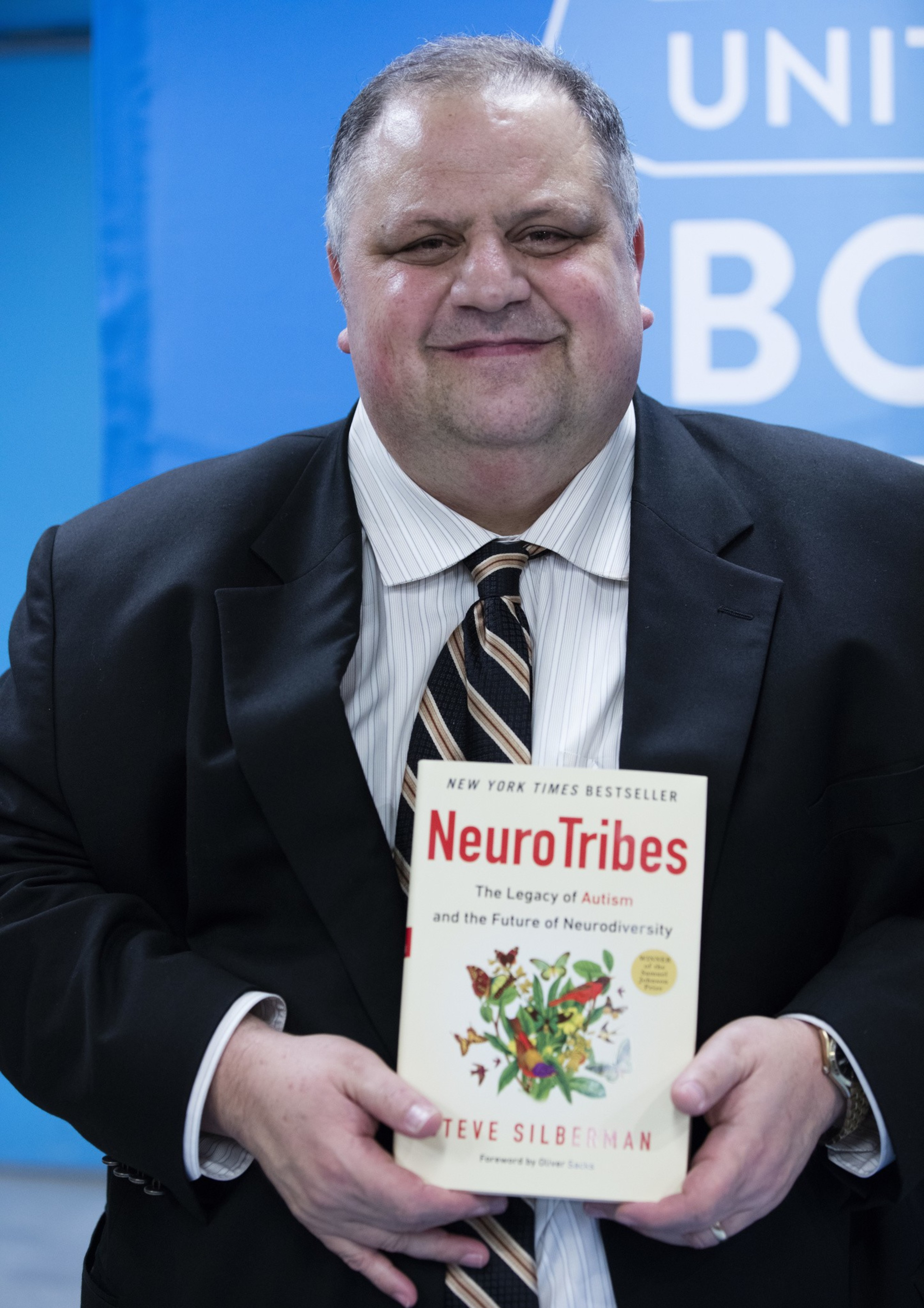 A man in a suit holds the book "NeuroTribes" by Steve Silberman, standing against a blue background with partial words visible.