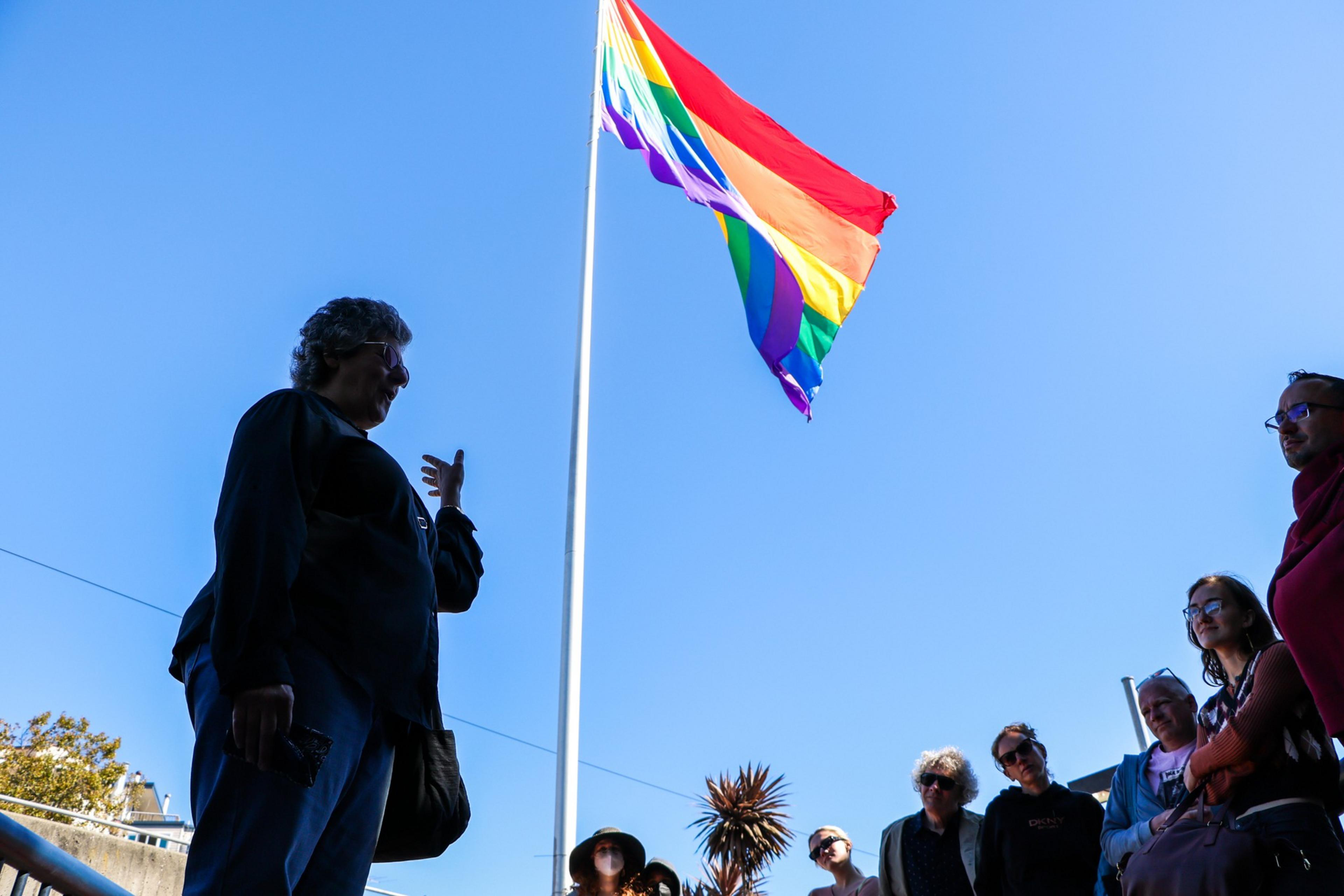A group of people stand outdoors under a bright blue sky, gathered near a tall pole flying a rainbow pride flag.