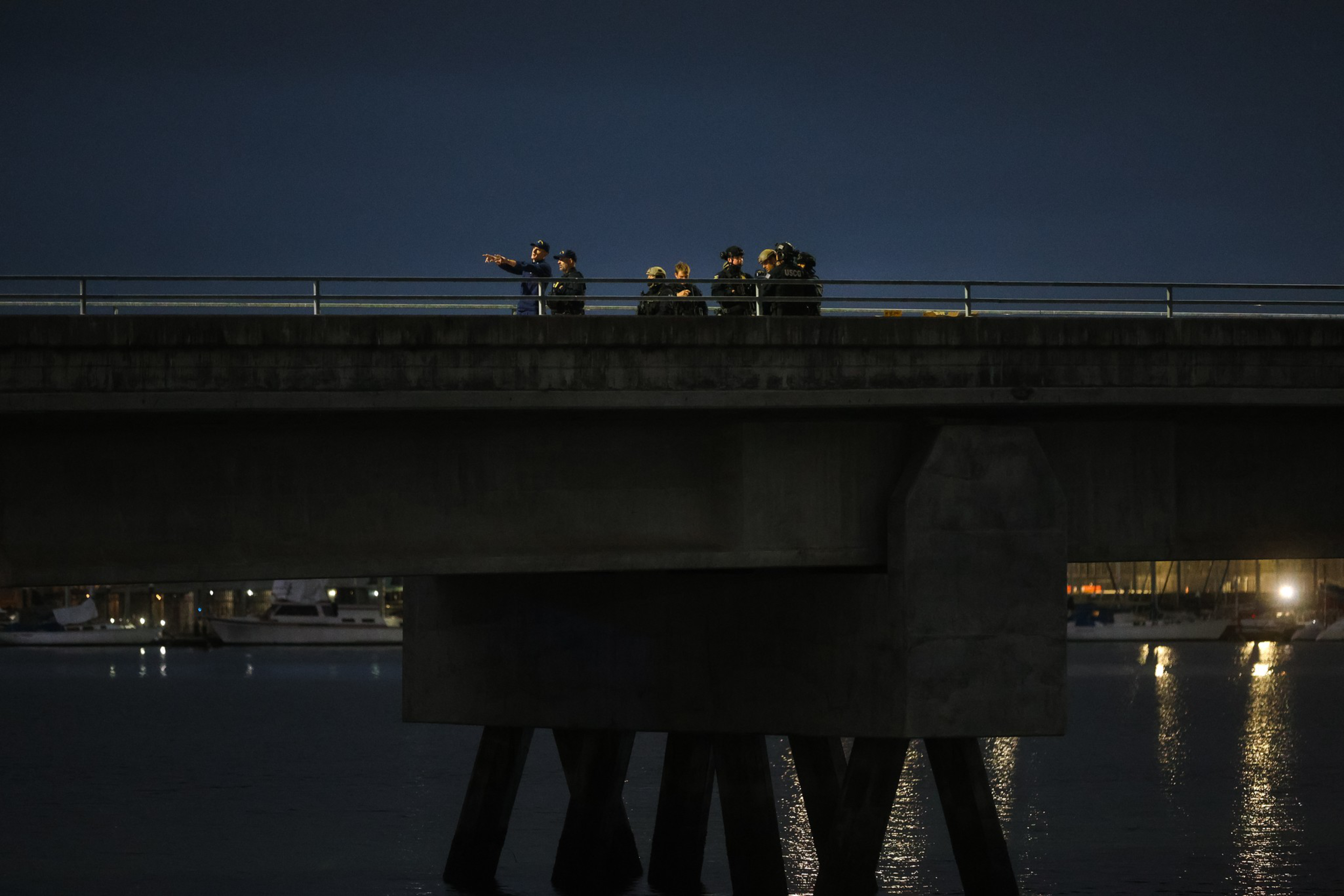 A group of people stand on a bridge at night, with one person pointing ahead, illuminated against a dark sky and water below.