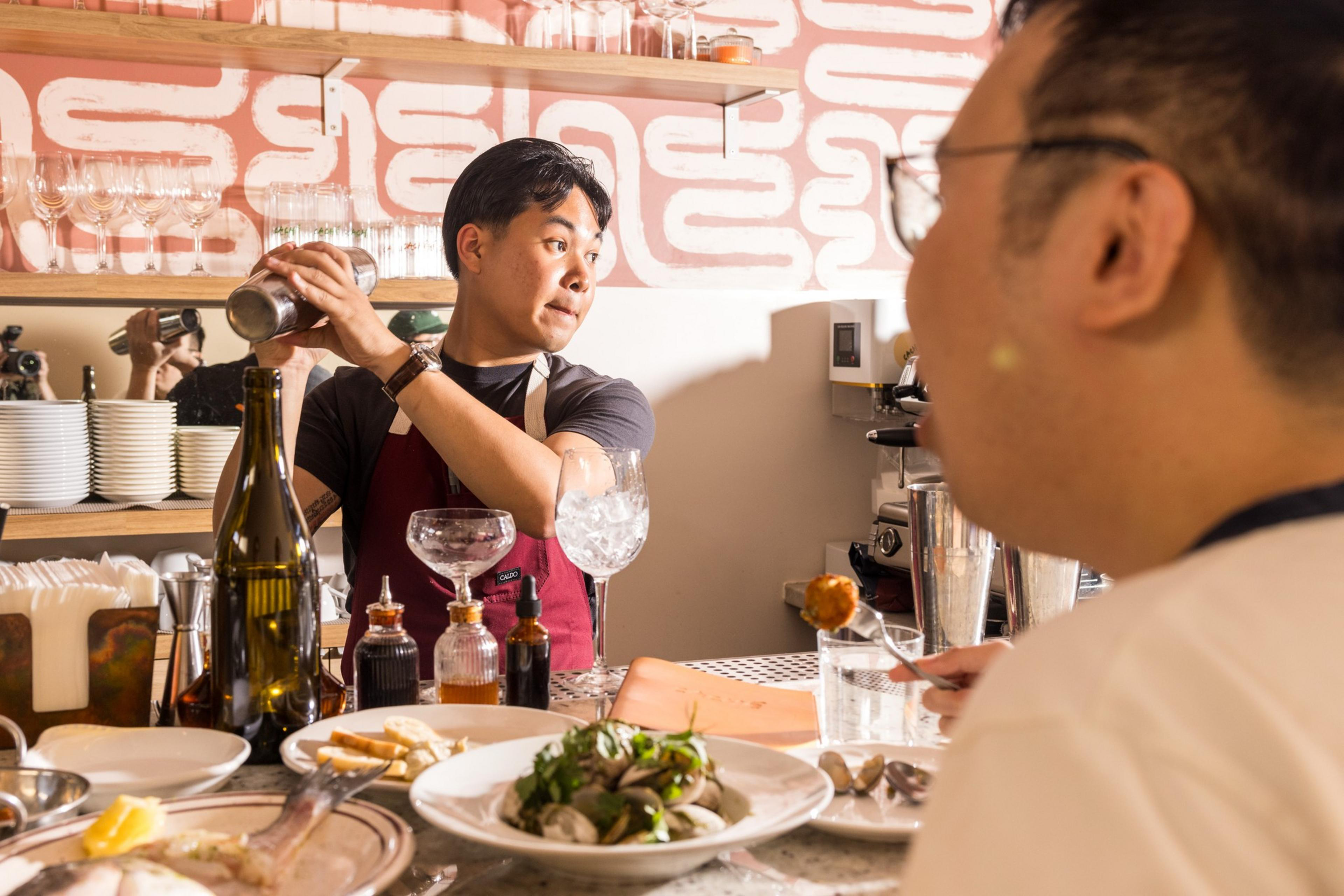 A bartender in an apron shakes a cocktail shaker while a person at the bar enjoys a meal, surrounded by dishes and bottles in a warmly lit setting.