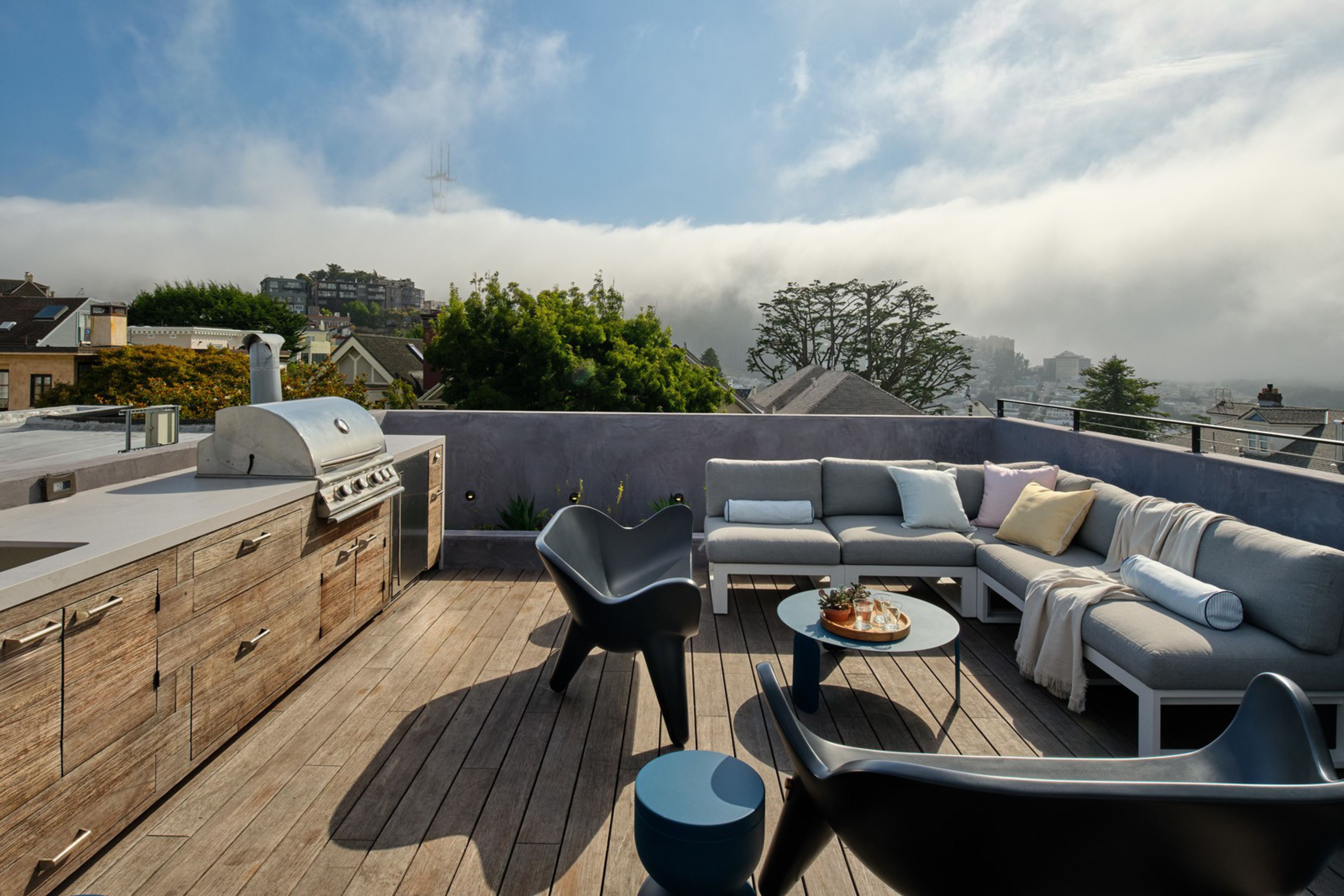 A rooftop patio features a wooden deck, built-in barbecue grill, gray sectional sofa with colorful pillows, black chairs, a round coffee table, and distant foggy hills.