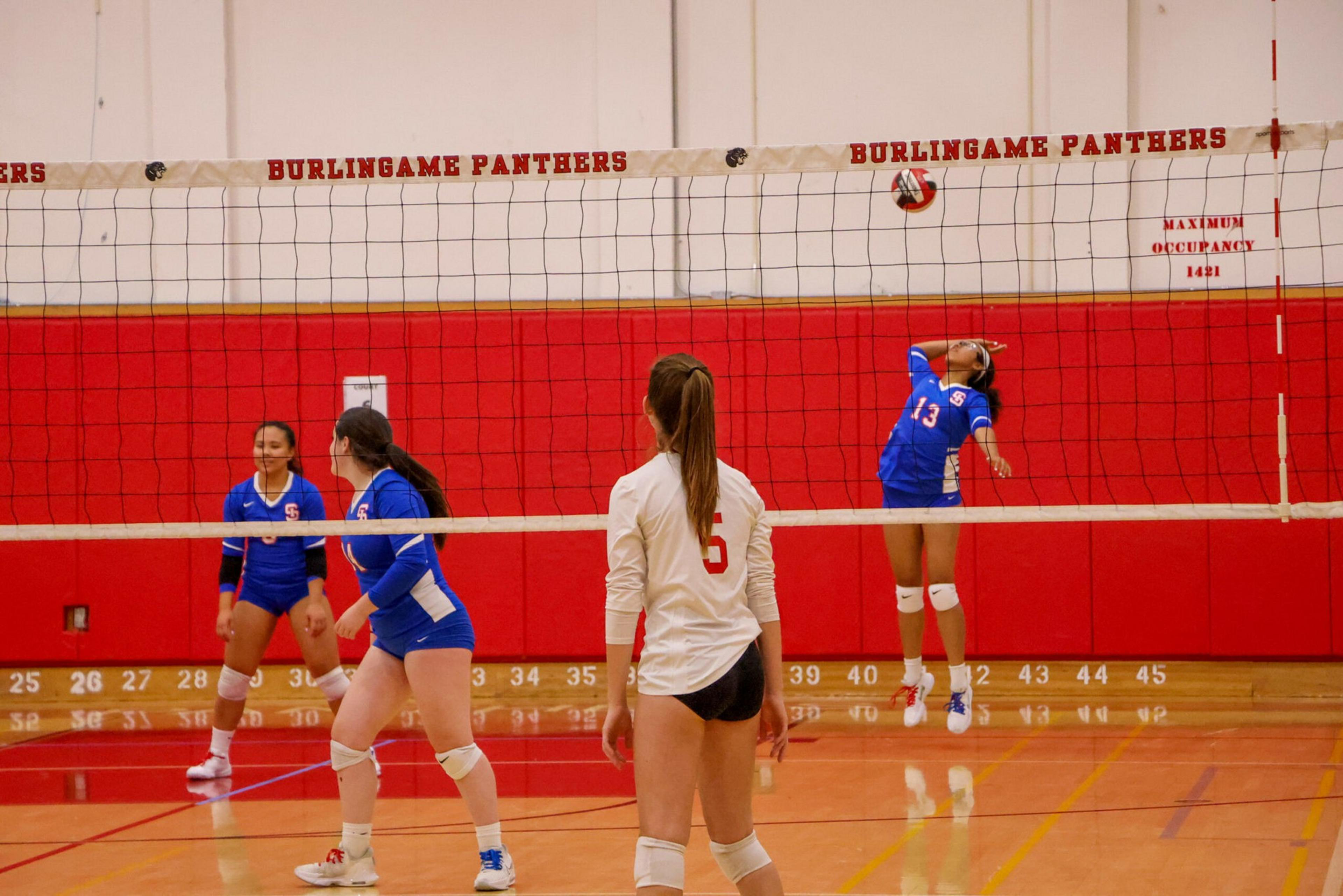 A volleyball player in blue jersey number 13 is mid-air hitting the ball while three other players, two in blue and one in white, watch.