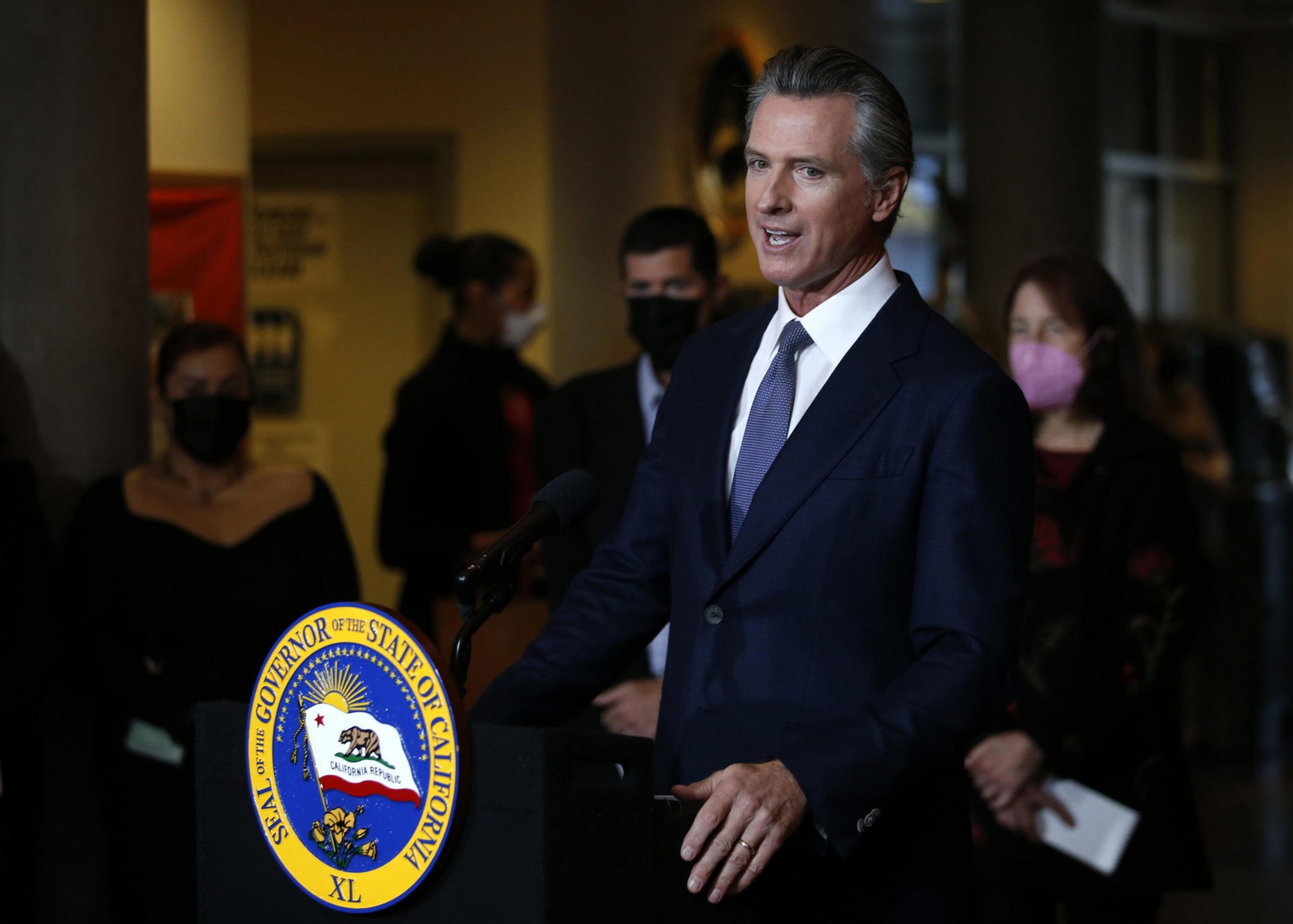 A man in a dark suit speaks at a podium with the California governor's seal, while people wearing masks stand behind him in a dimly lit room.
