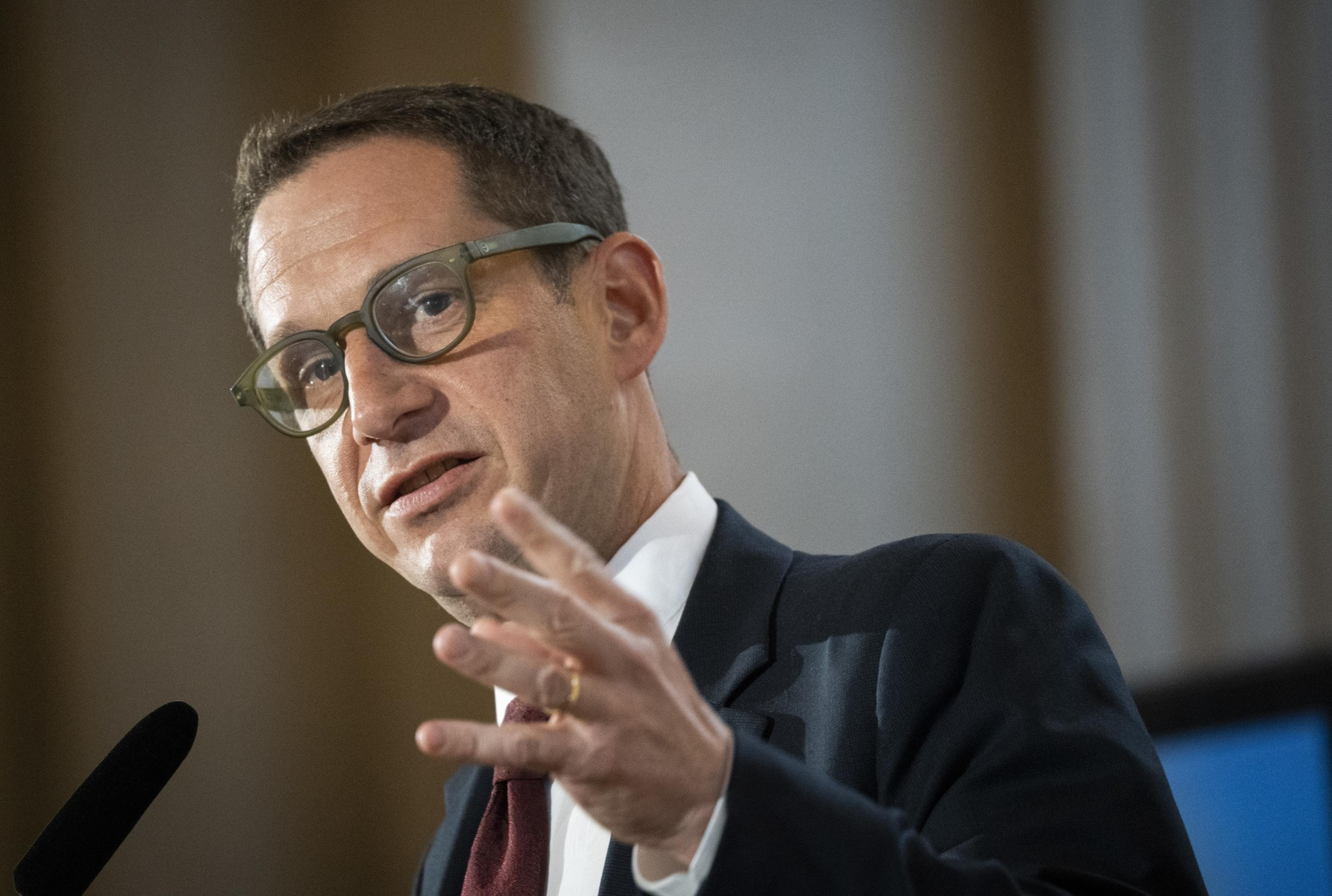 A person with glasses is speaking, gesturing with their hand. They wear a suit and tie, and there is a microphone visible. The background is blurred.