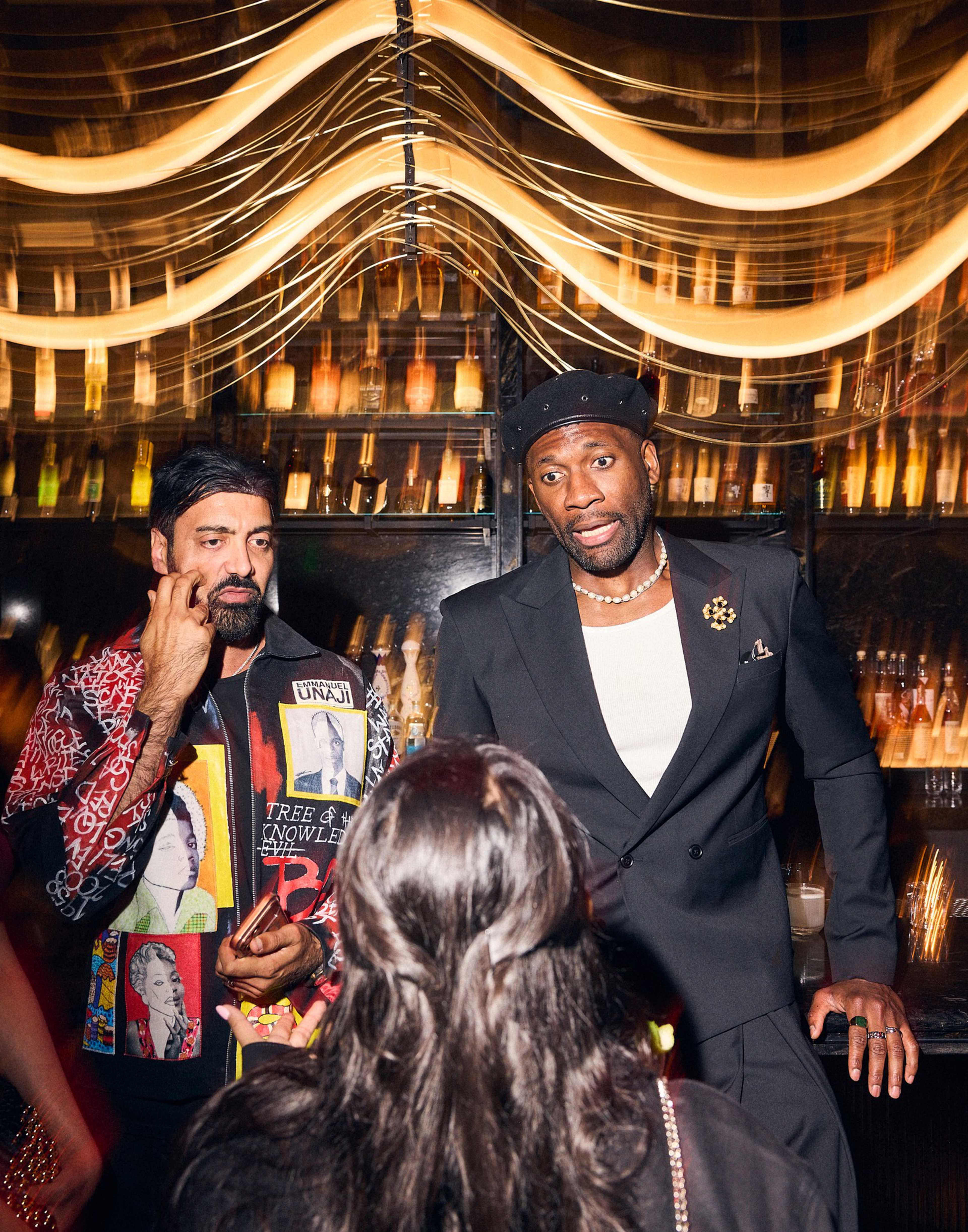 Two men stand behind a bar with illuminated bottles; one wears a patterned jacket and the other a black suit and beret, while a woman faces them.