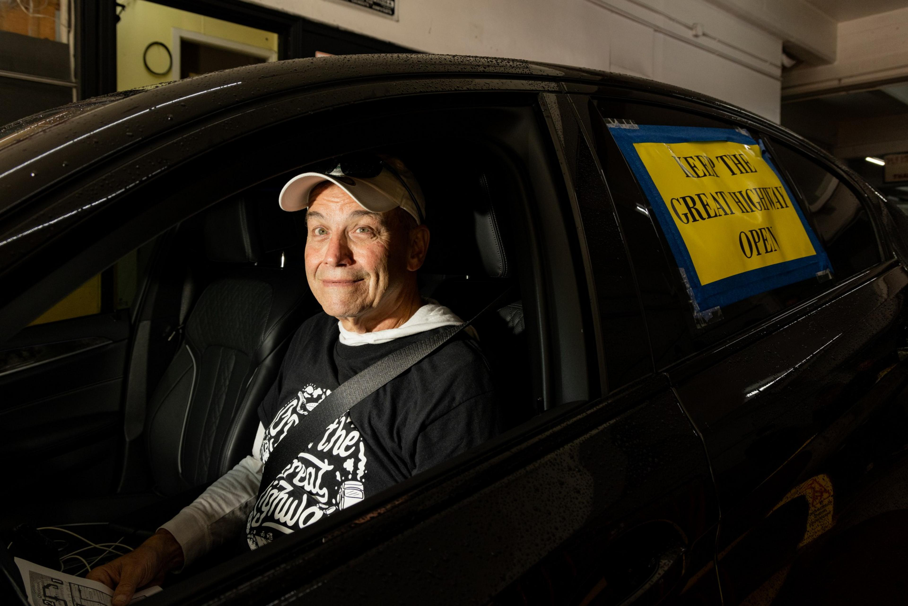A man wearing a white baseball cap and a black shirt sits in the driver's seat of a car. The car window has a yellow sign reading &quot;KEEP THE GREAT HIGHWAY OPEN.&quot;