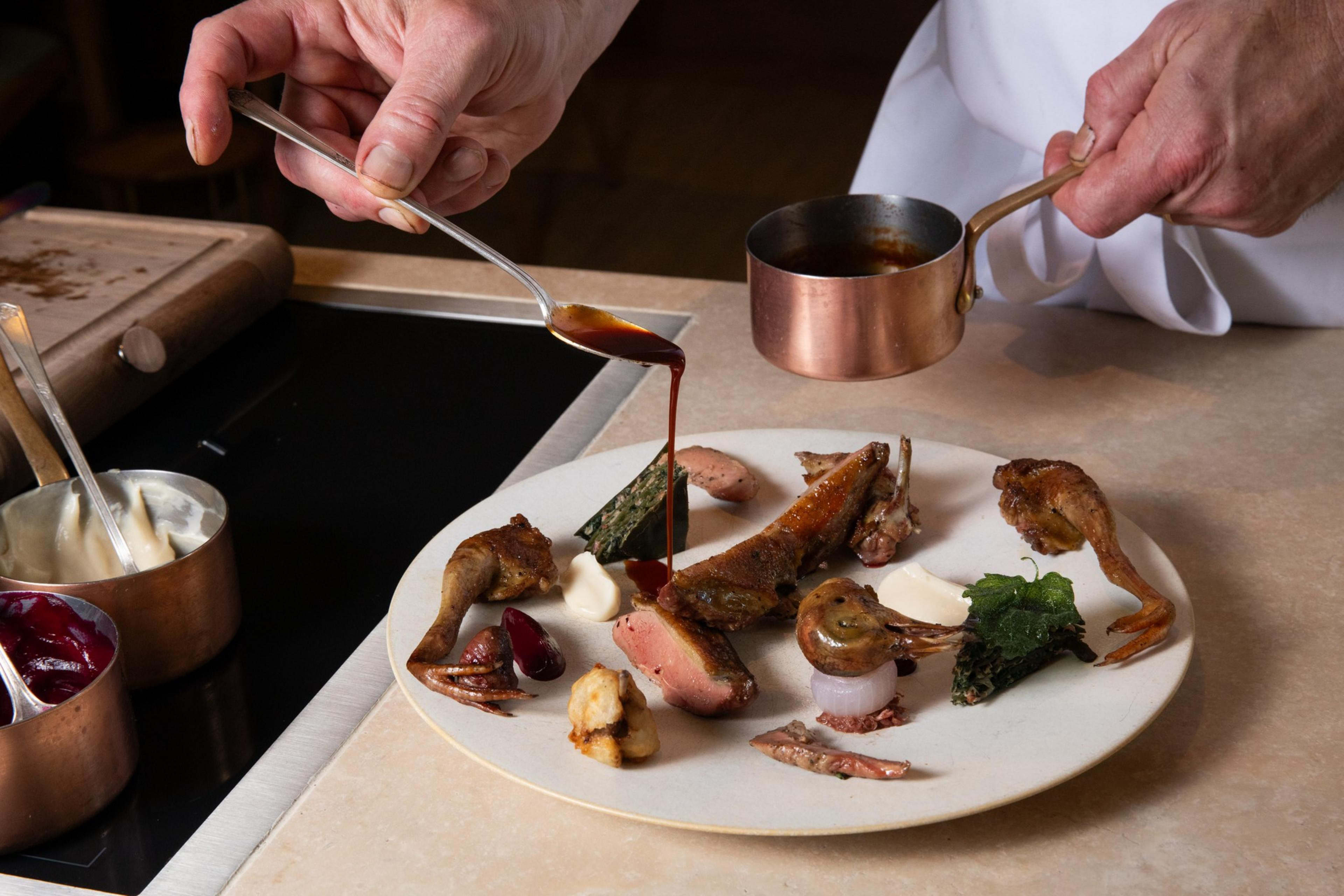 A chef is artfully plating a gourmet dish. The plate has various meats, sauces, and garnishes. The chef pours sauce from a copper pot onto the dish.