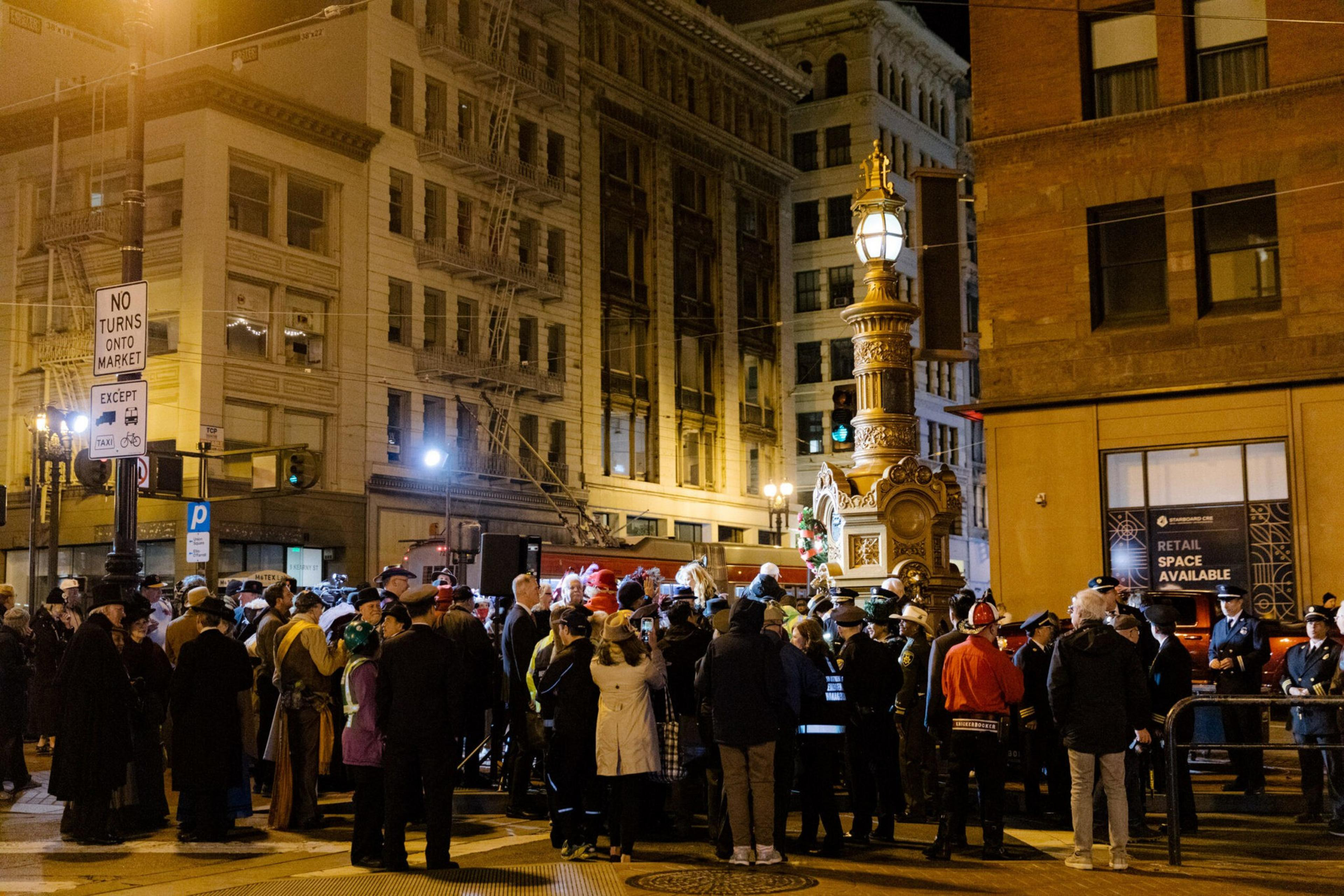 A crowd gathers at night around a decorative street lamp in an urban area with historic buildings and a "No Turns Onto Market" sign nearby.
