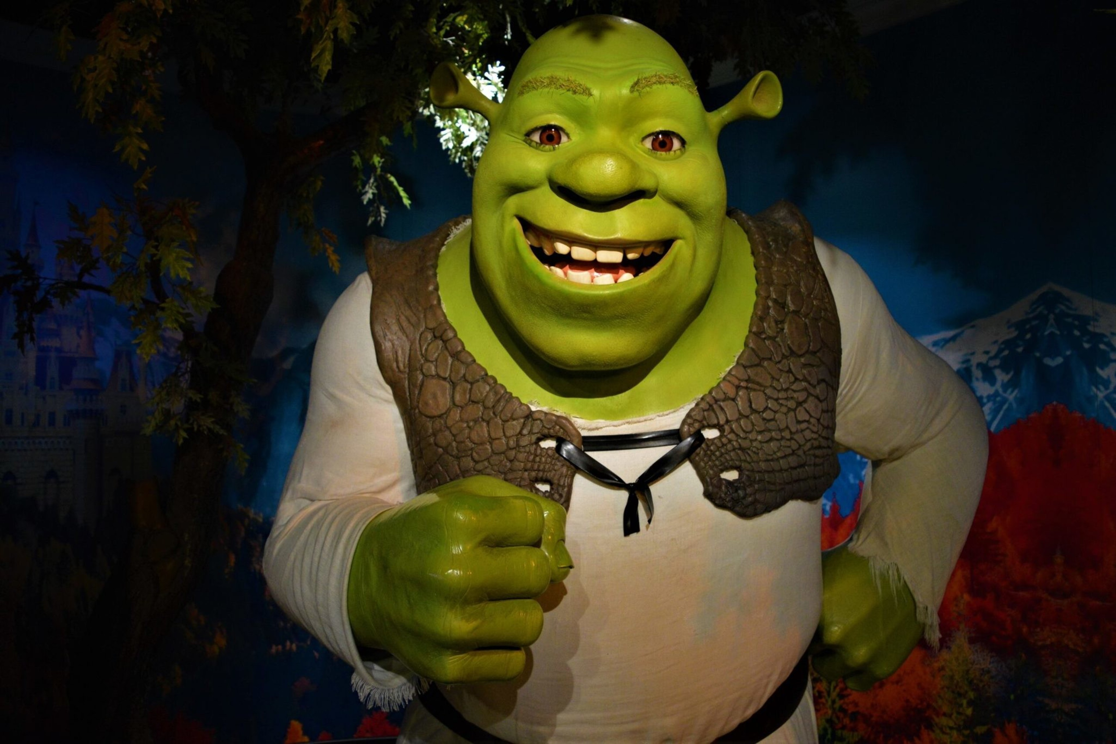 A smiling green ogre with round ears and wearing a white shirt and vest stands with one fist raised and the other on its hip.
