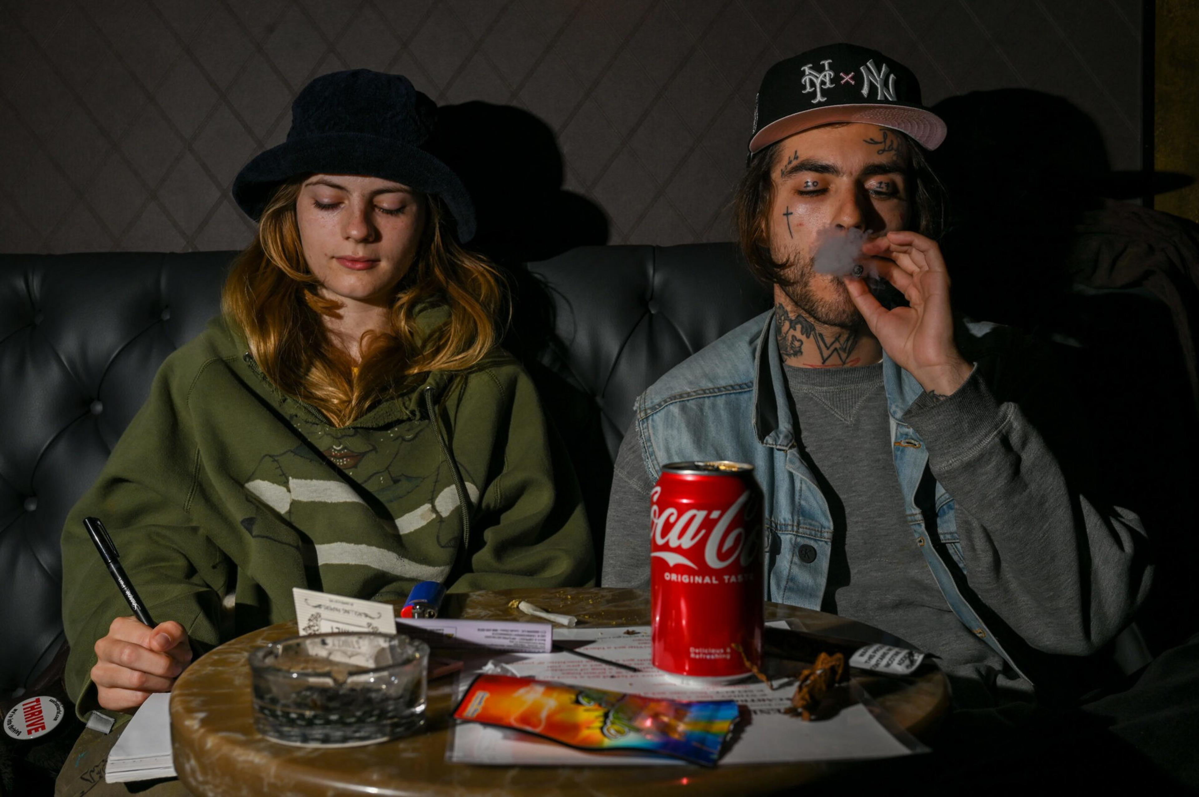 A young woman in a green hoodie writes while a tattooed man in a cap exhales smoke next to her; a Coca-Cola can and various items sit on the table.