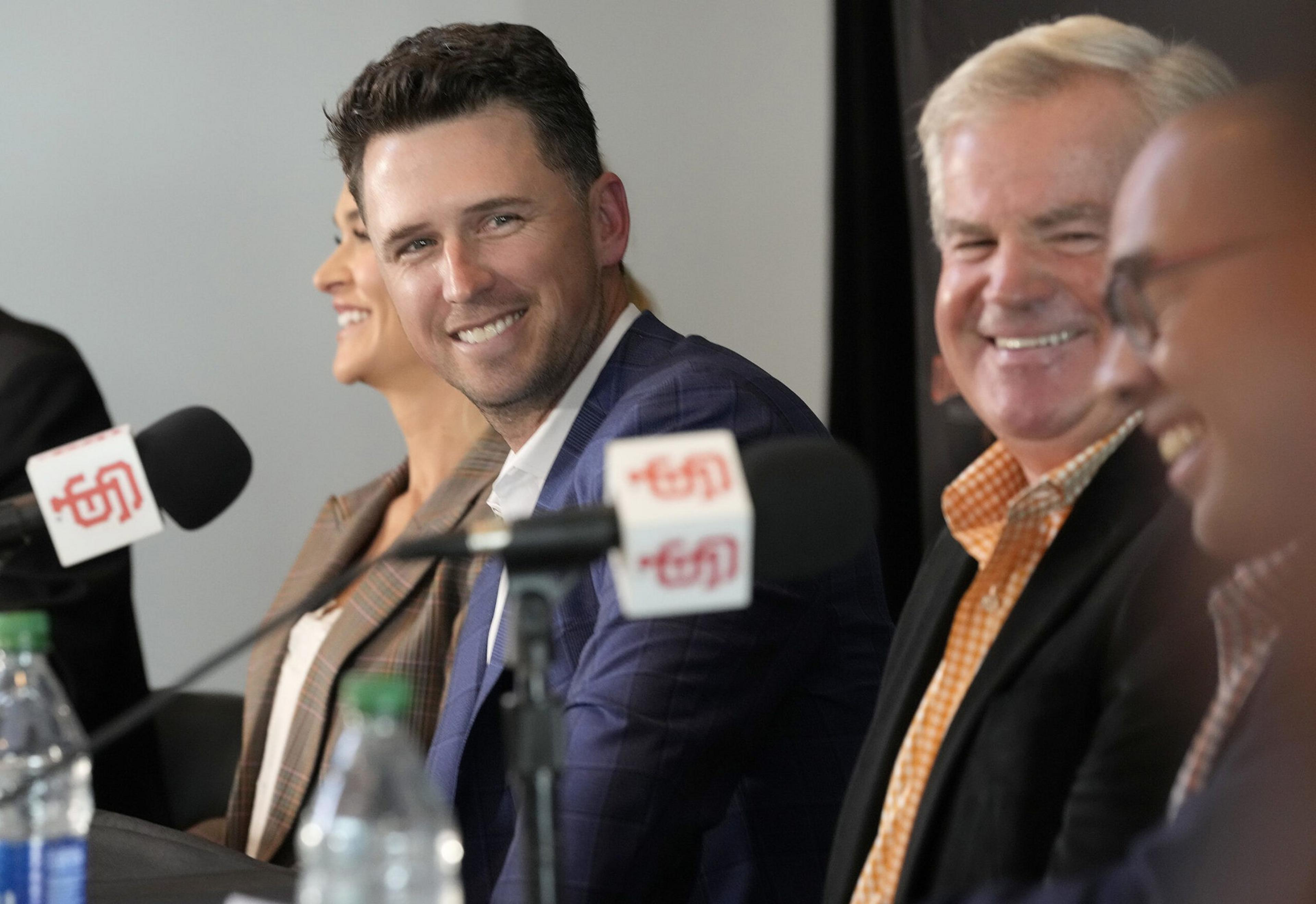 Several people, including a smiling man in a blue suit, sit at a table with microphones and water bottles, appearing to be at a press conference or panel discussion.