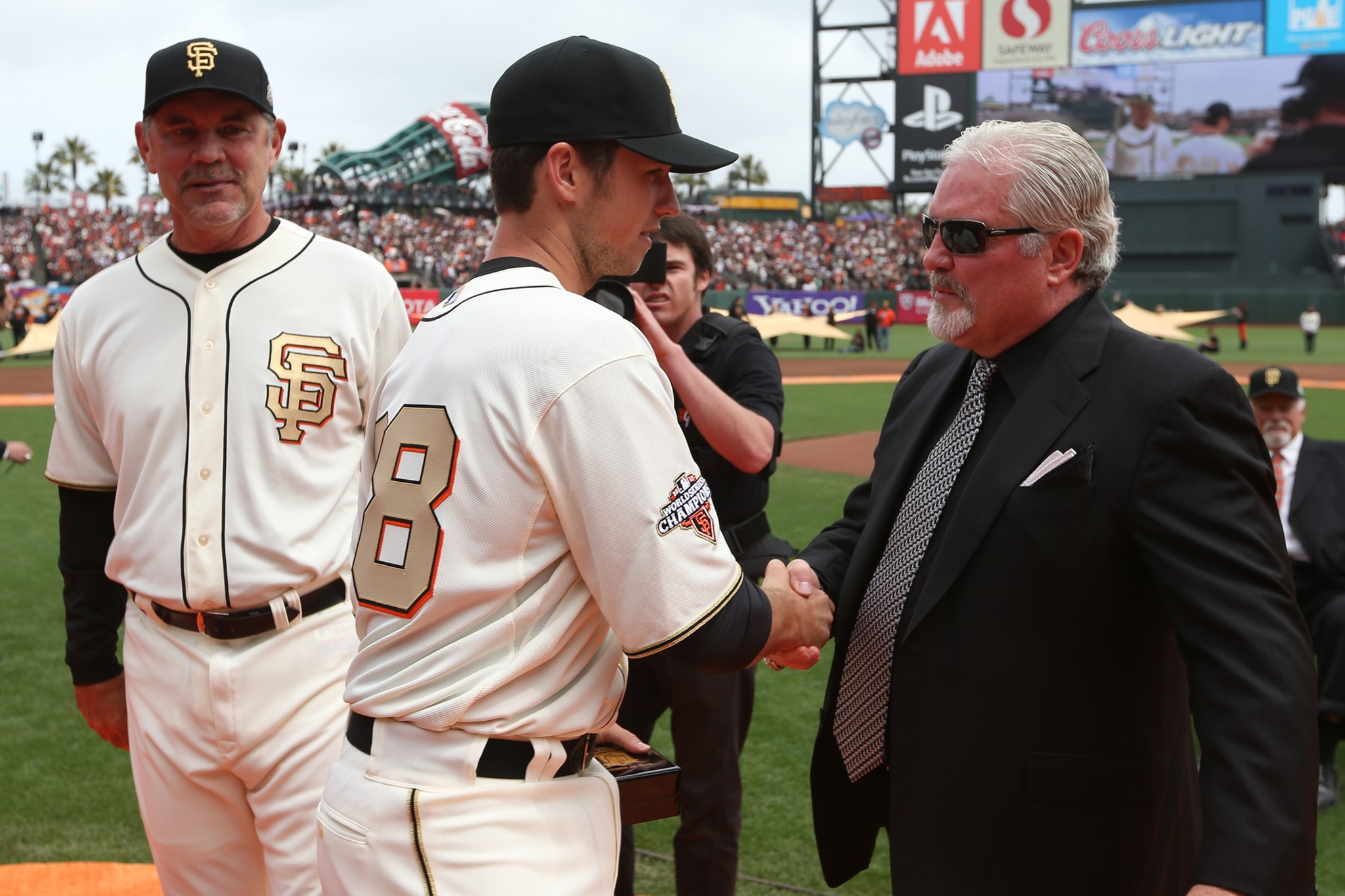 A baseball player in a white Giants uniform shakes hands with a man in a suit and sunglasses. Another person in a Giants uniform stands nearby.