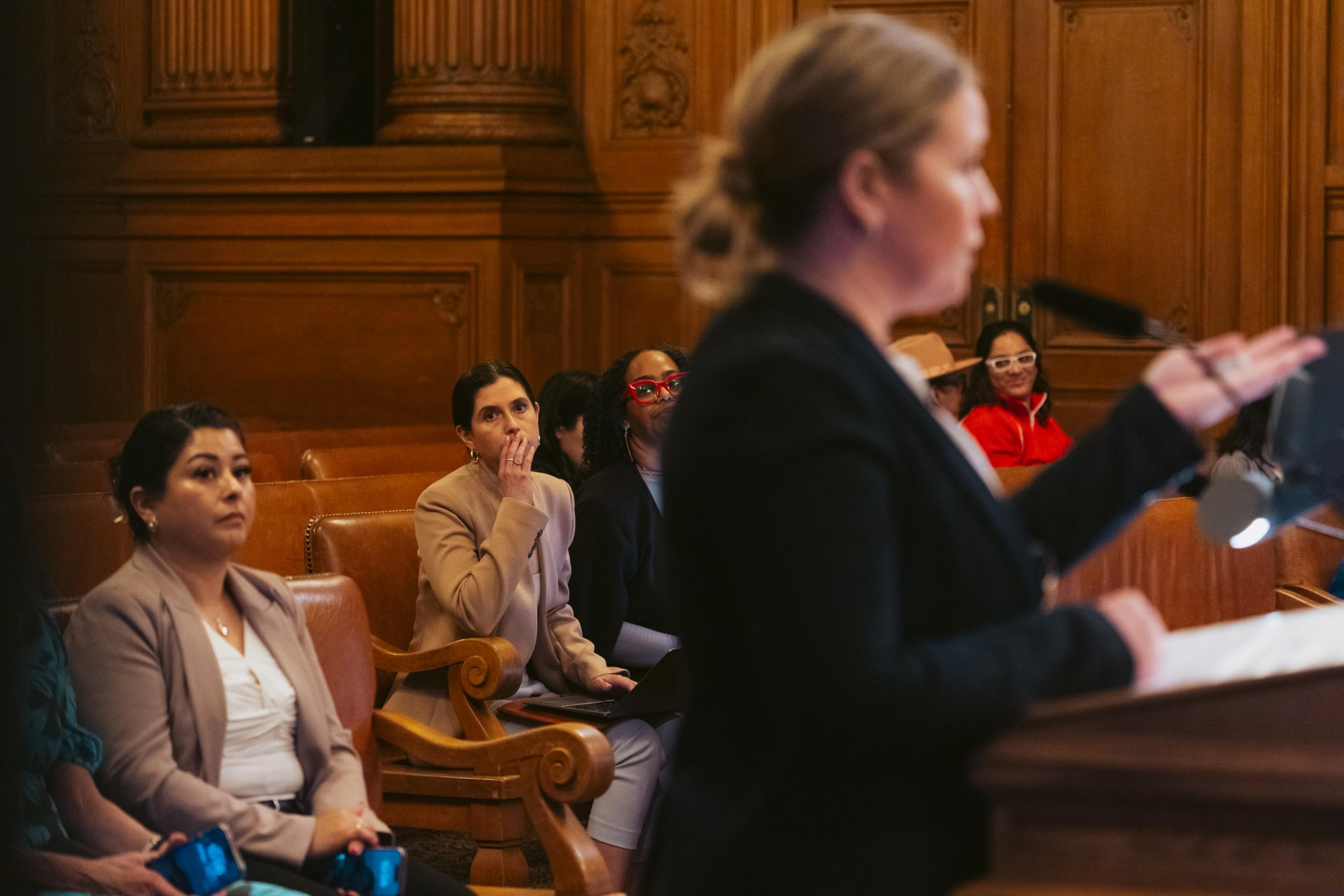 An audience of women listen—one holds her hands over her mouth—as a police captain speaks at a podium about sexual assault.