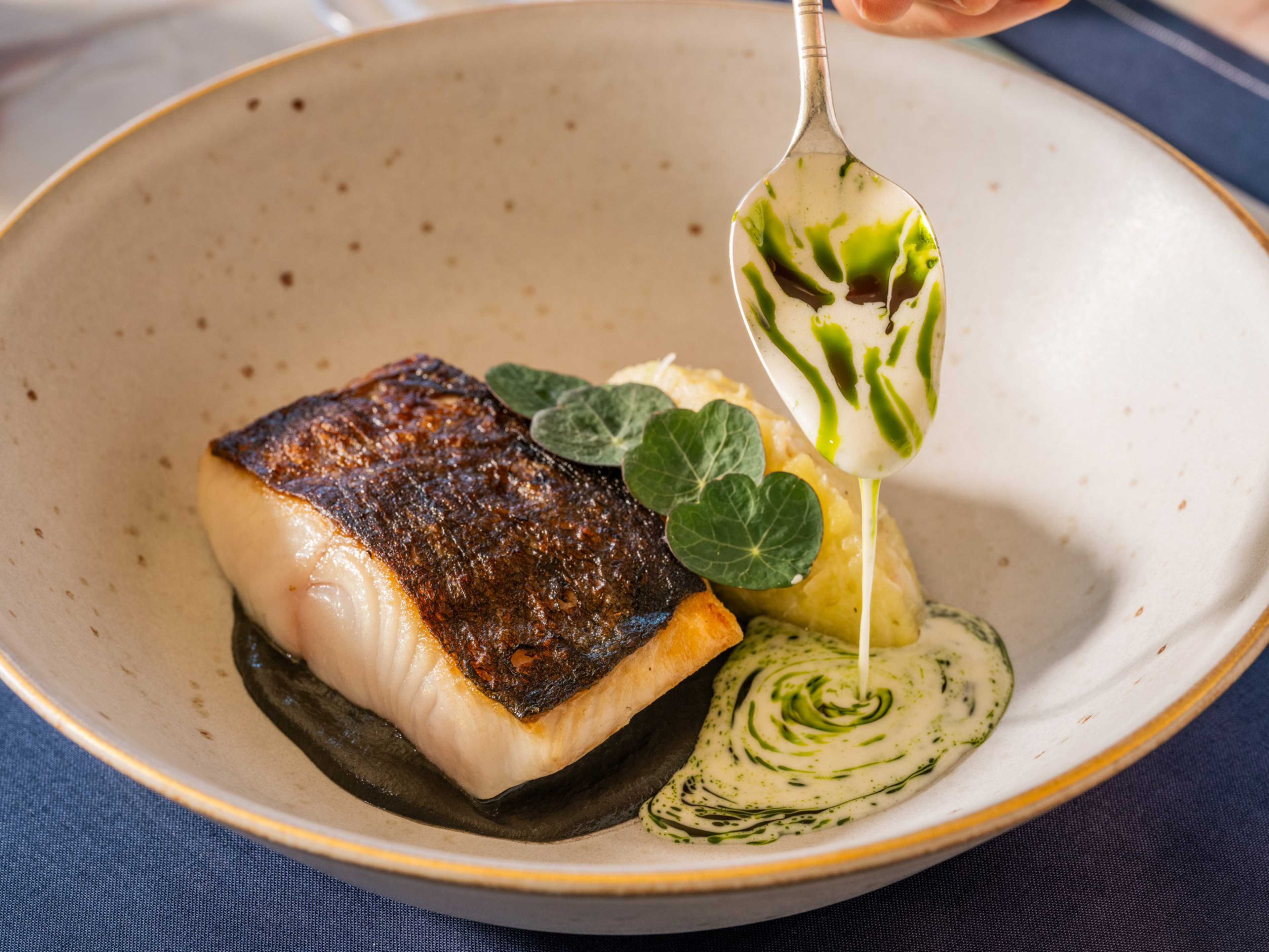 A seared fish fillet with crispy skin sits on a dark sauce, accompanied by mashed potatoes, green leaves, and a creamy green herb sauce being drizzled.