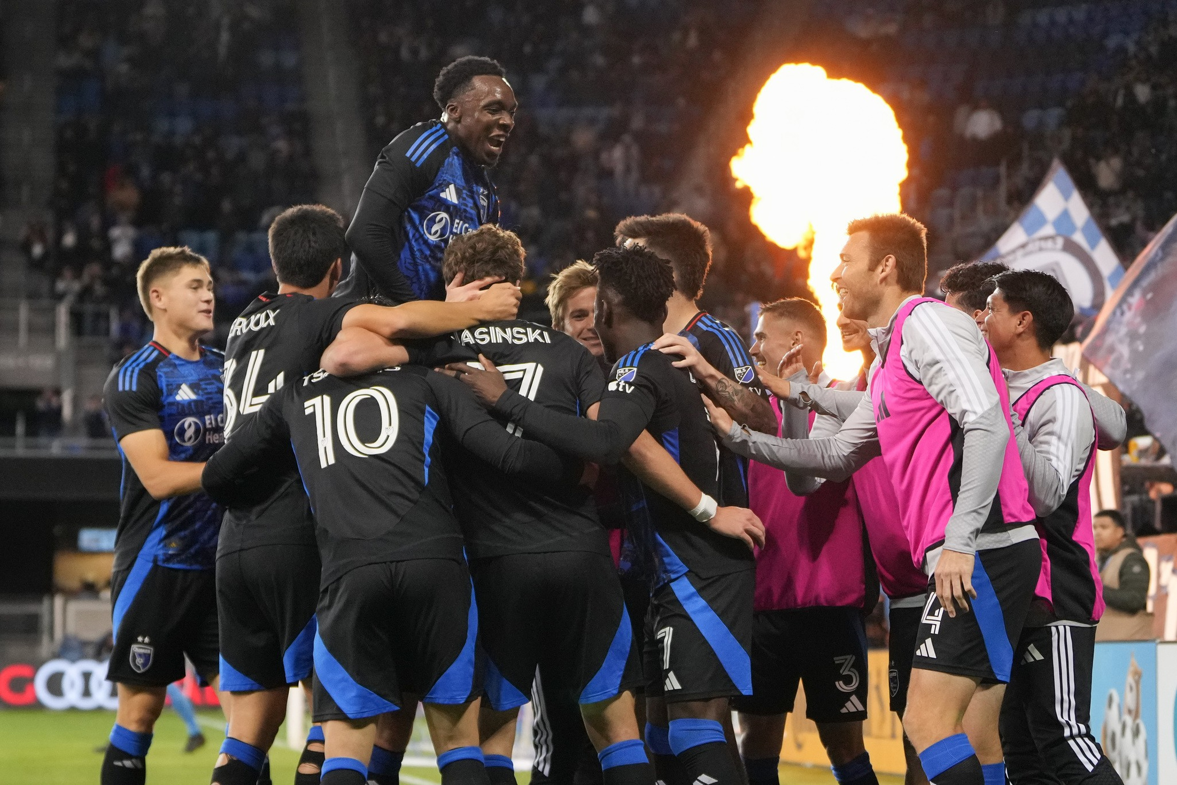 Soccer players in black and blue uniforms celebrate together with one player lifted on teammates’ shoulders, and a large flame bursts in the background.