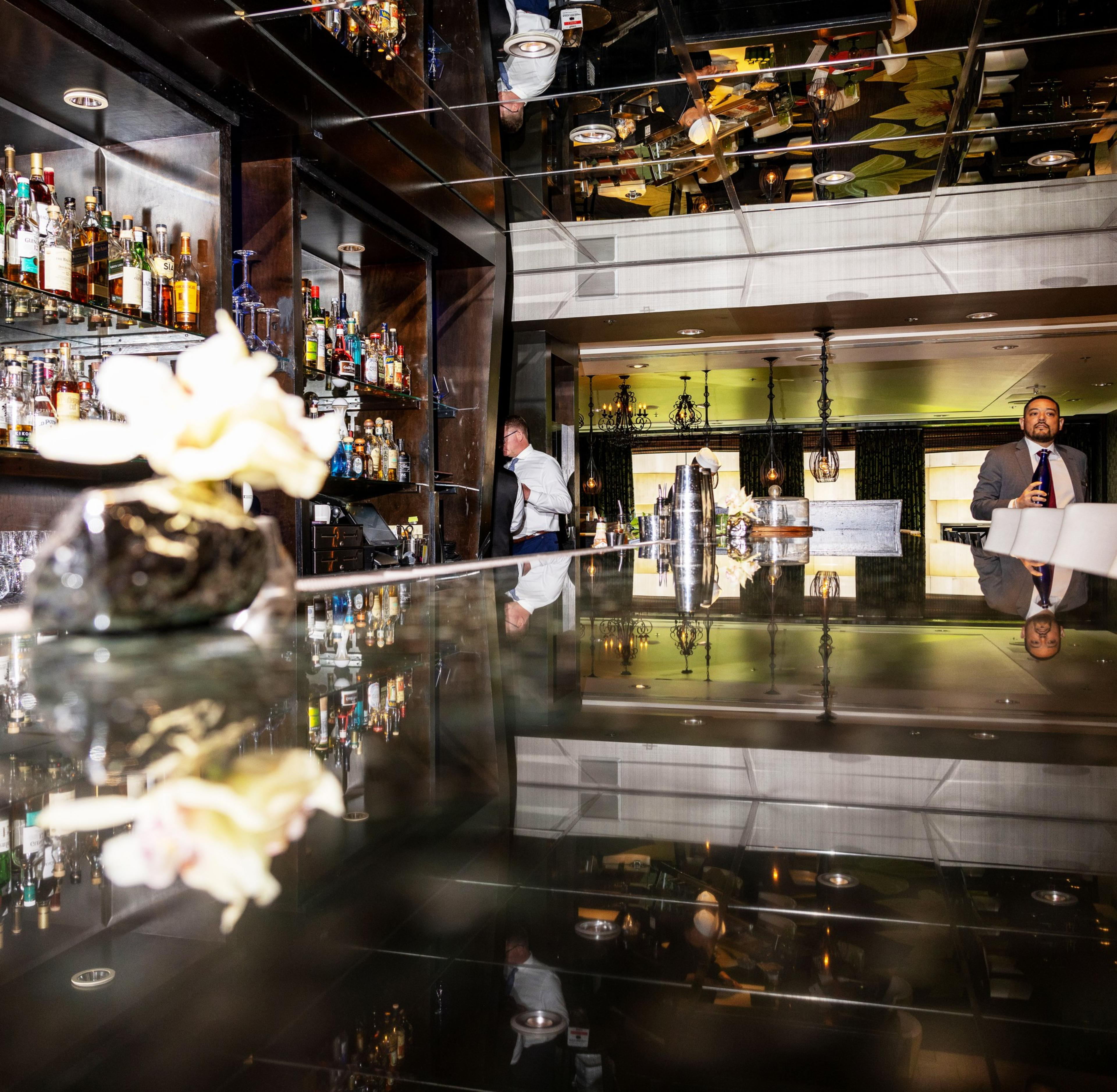 The image shows a sleek, modern bar with mirrored surfaces reflecting bottles and lights. A bartender is working, while a suited man with a drink stands nearby.