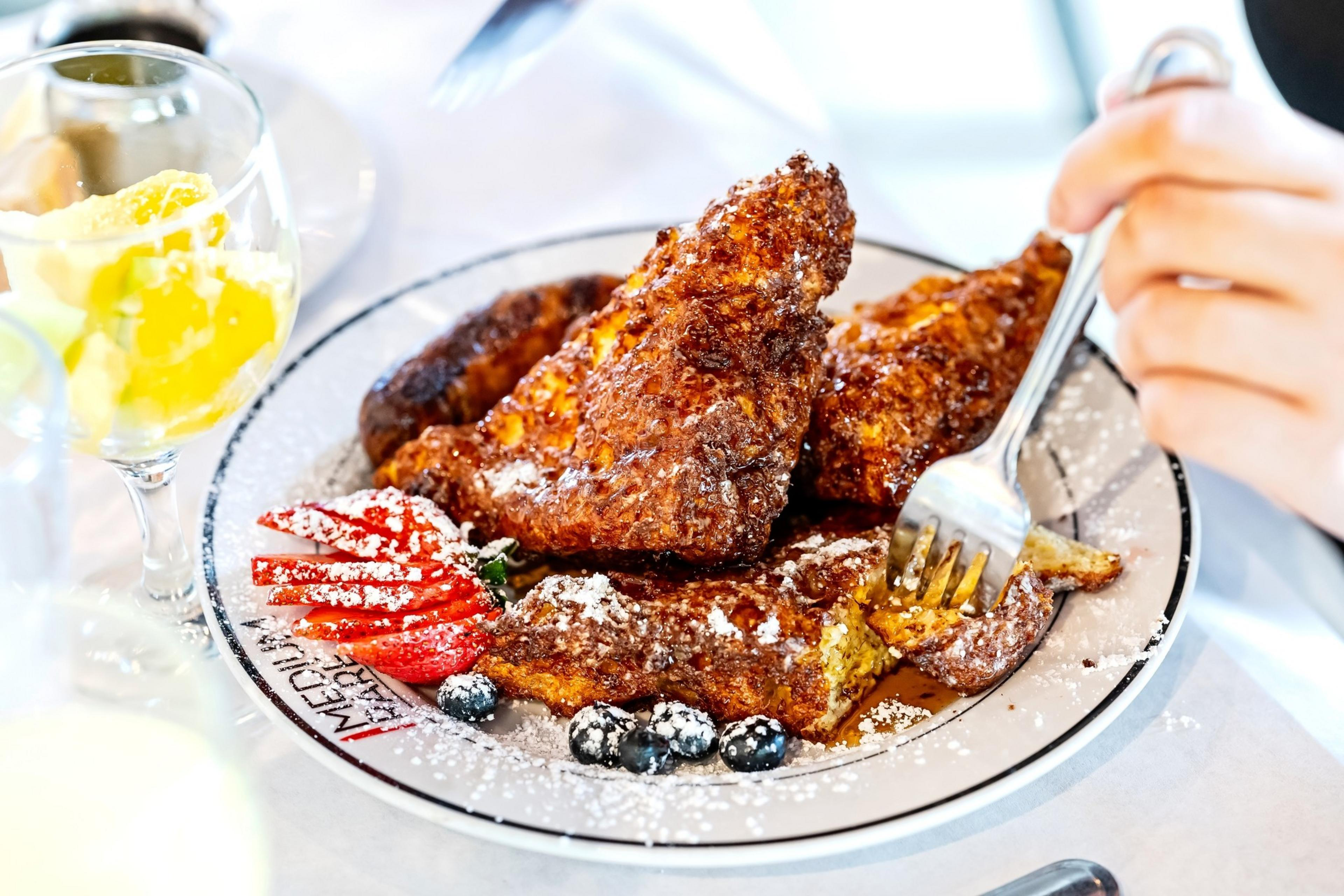 A person's hand holds a fork over a plate bearing several slices of rich looking French toast with sliced strawberries and a few blueberries.