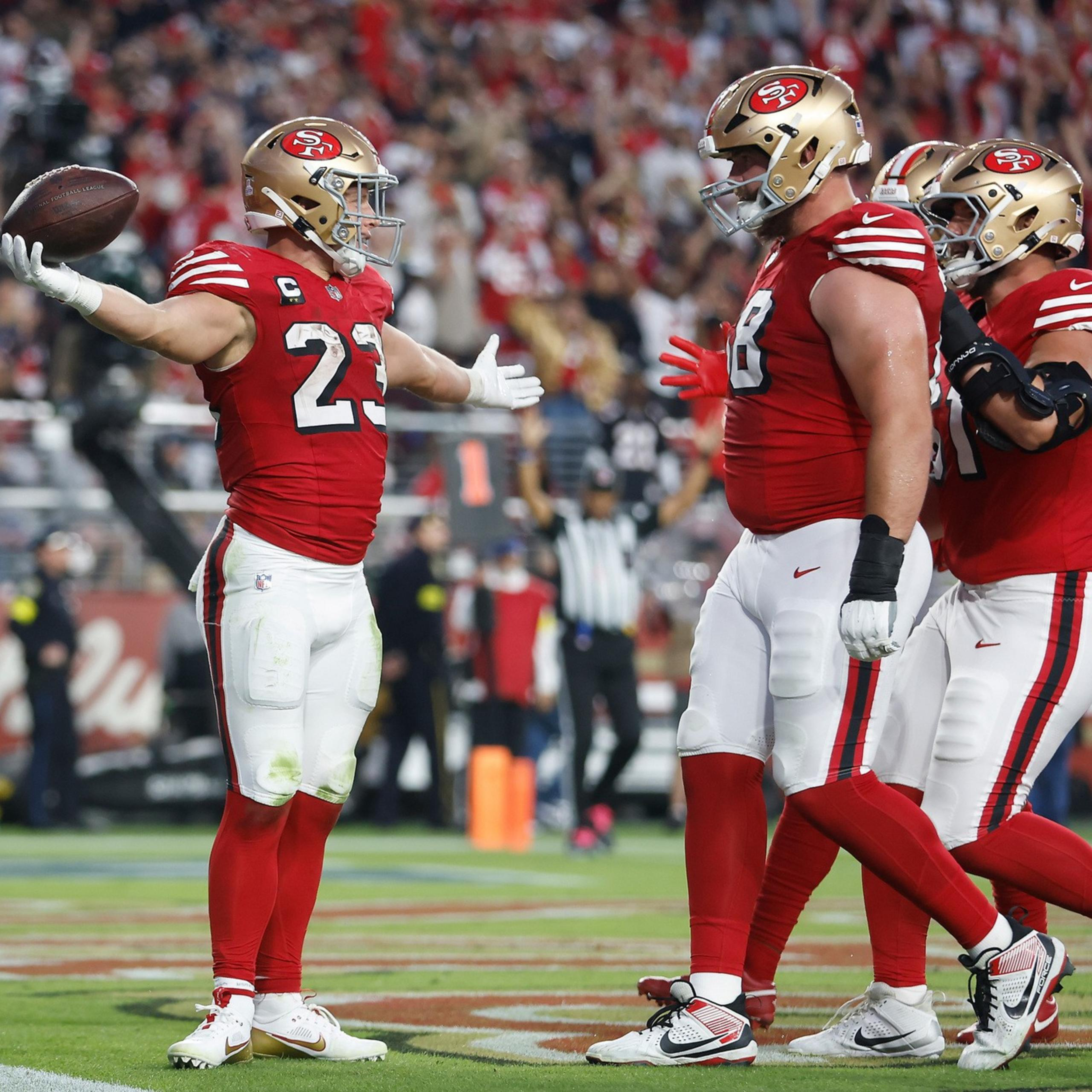 Three San Francisco 49ers football players in red and white uniforms celebrate on the field as one holds a football and a cameraman films them.