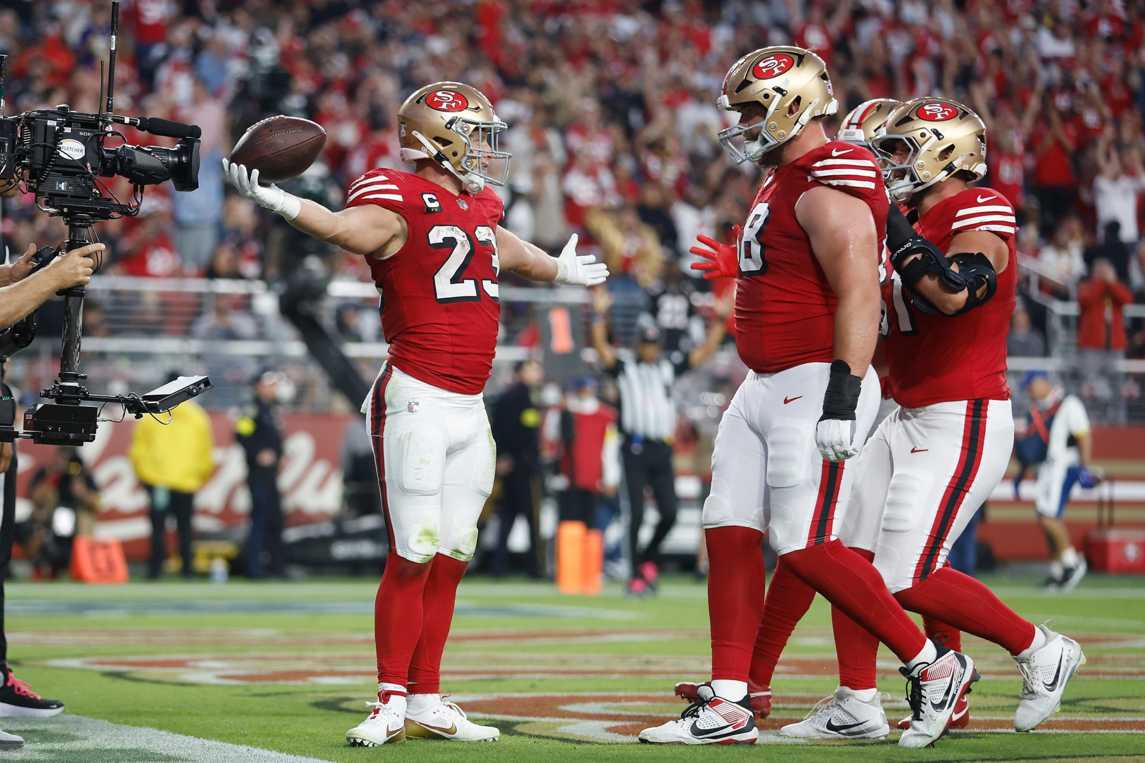 Three San Francisco 49ers football players in red and white uniforms celebrate on the field as one holds a football and a cameraman films them.