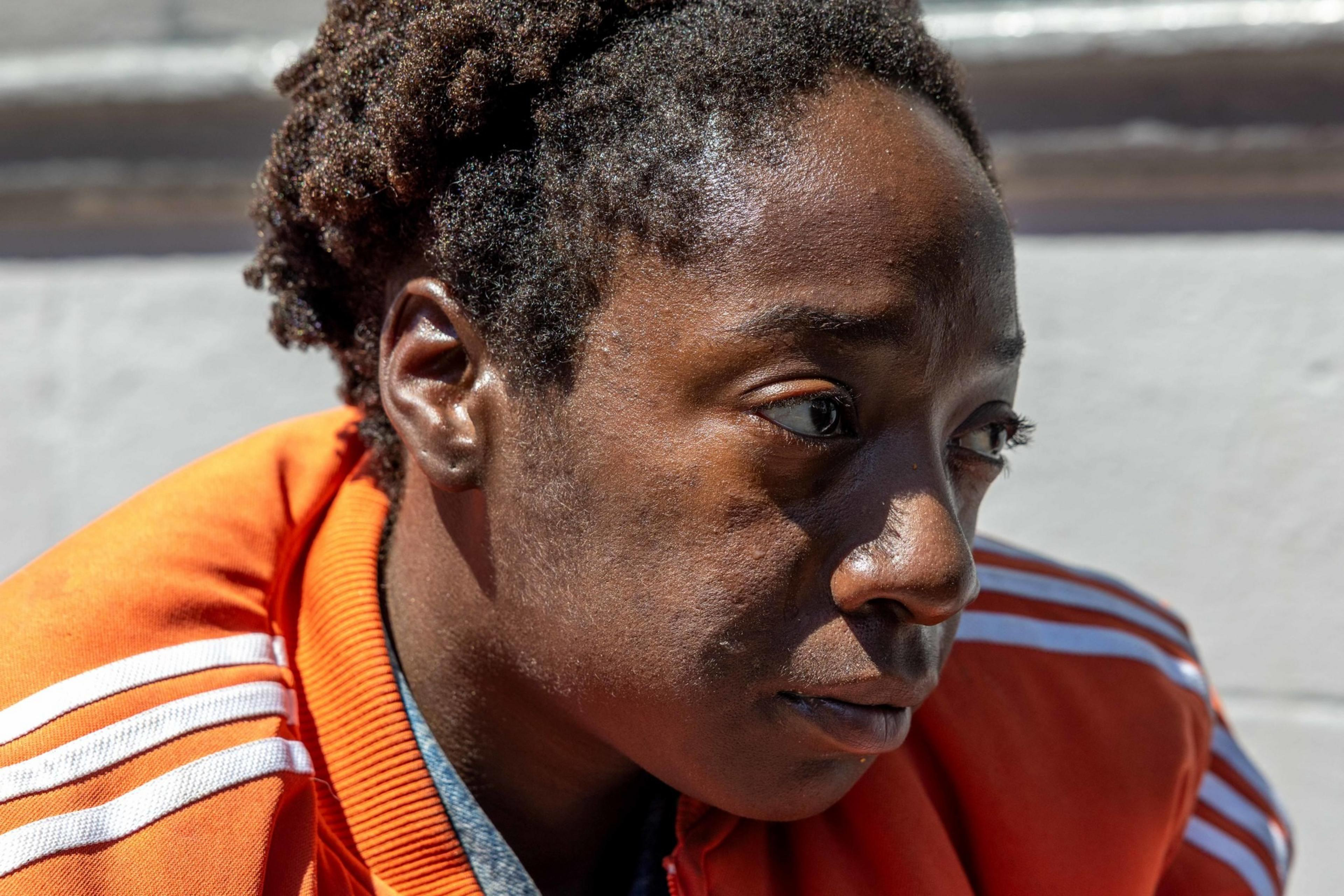 A person with short, curly hair is wearing an orange jacket with white stripes. The image focuses on their face, showing a thoughtful expression.