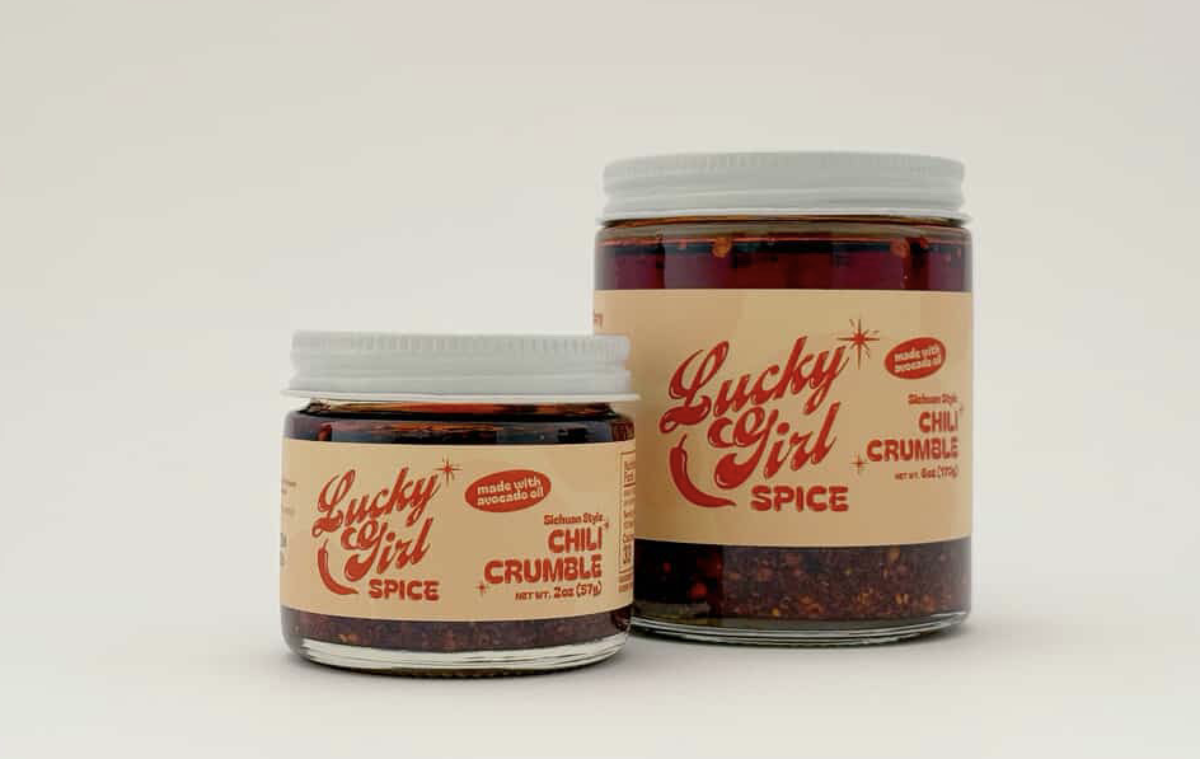 Two jars of Lucky Girl Spice Sichuan Style Chili Crumble with white lids and beige labels, featuring red text and a chili pepper graphic, sit side by side.