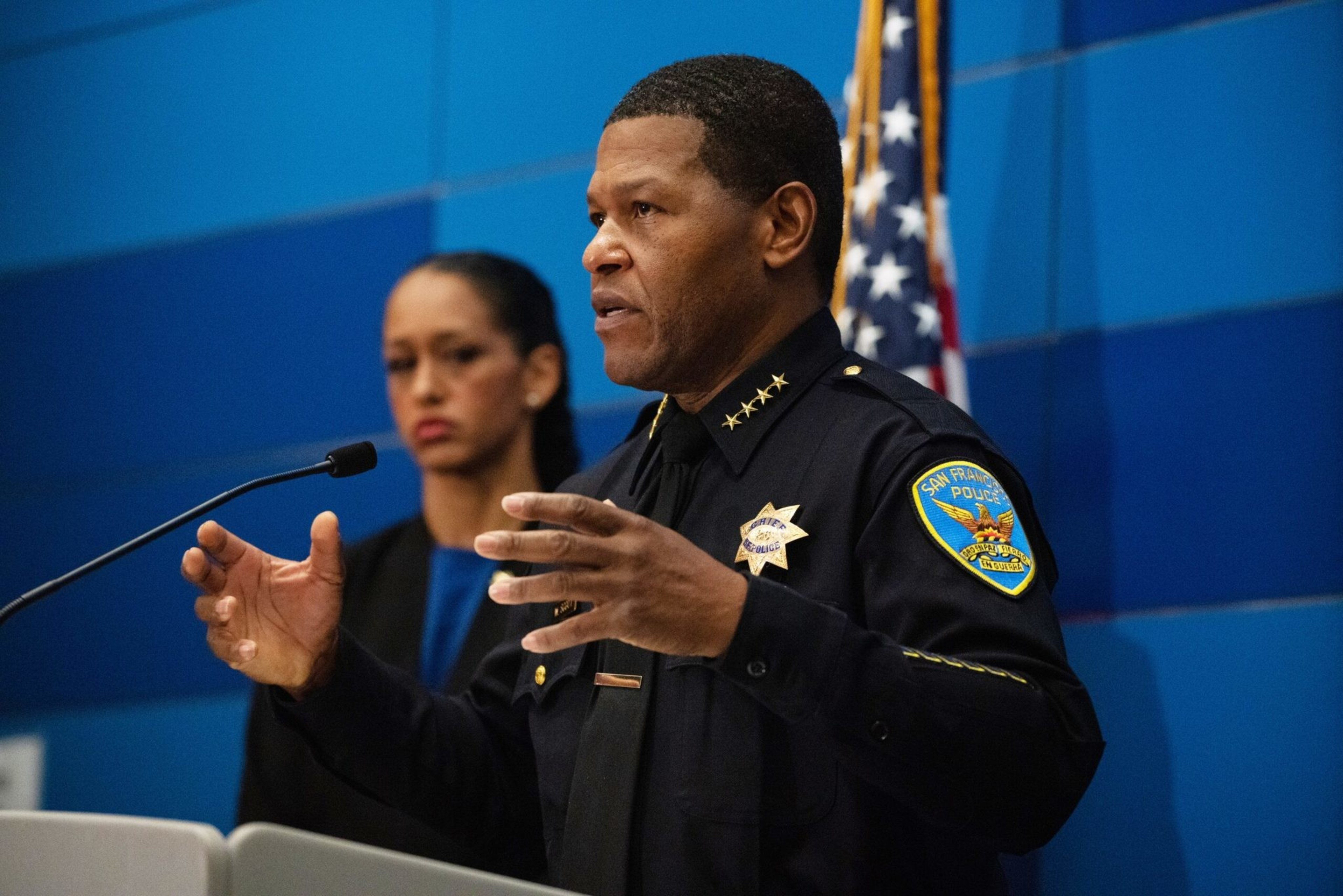 A man in a San Francisco police uniform speaks with hand gestures, while a woman stands behind him near a microphone and an American flag.