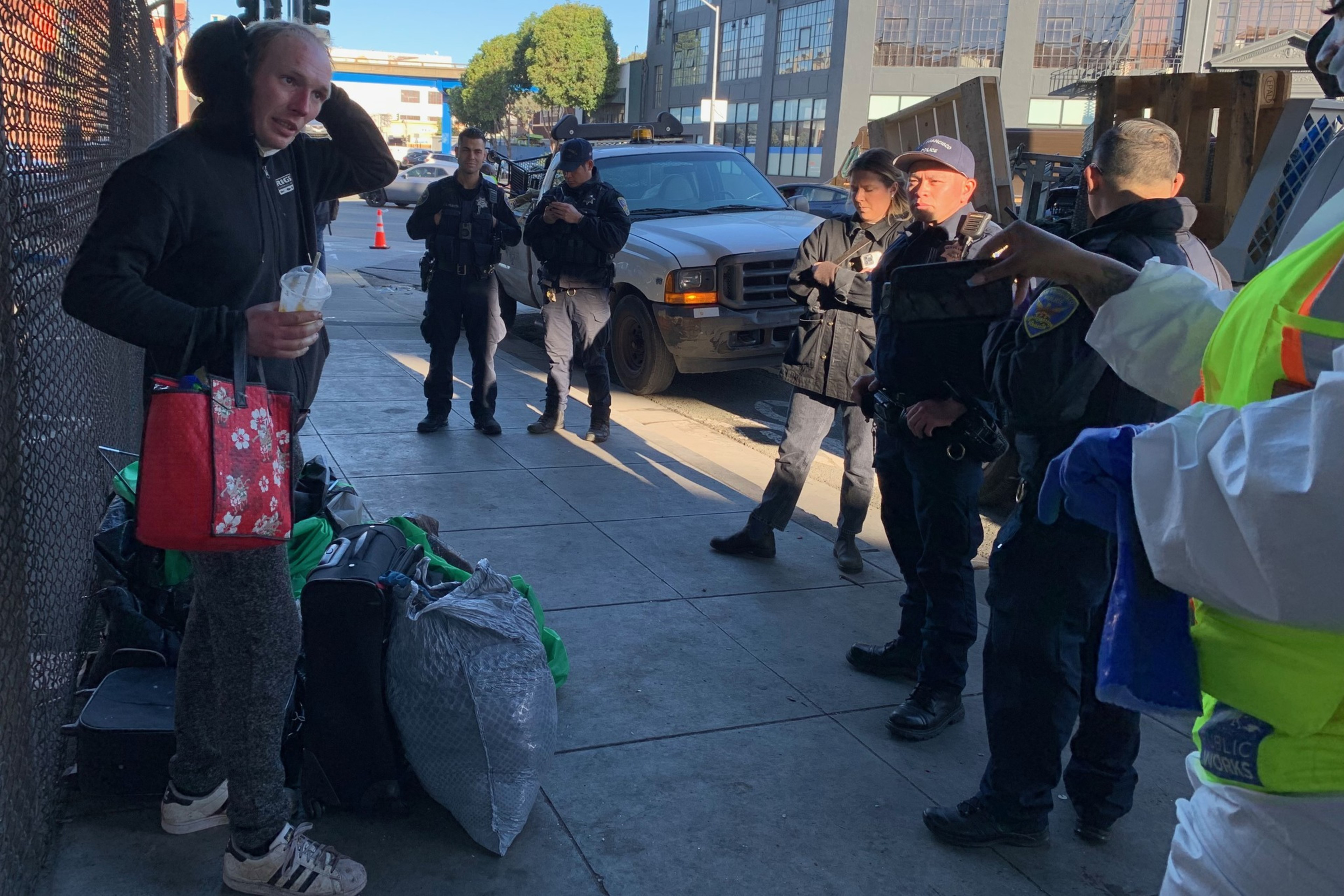 A person stands by belongings on a sidewalk, surrounded by police officers, with a vehicle in the background.