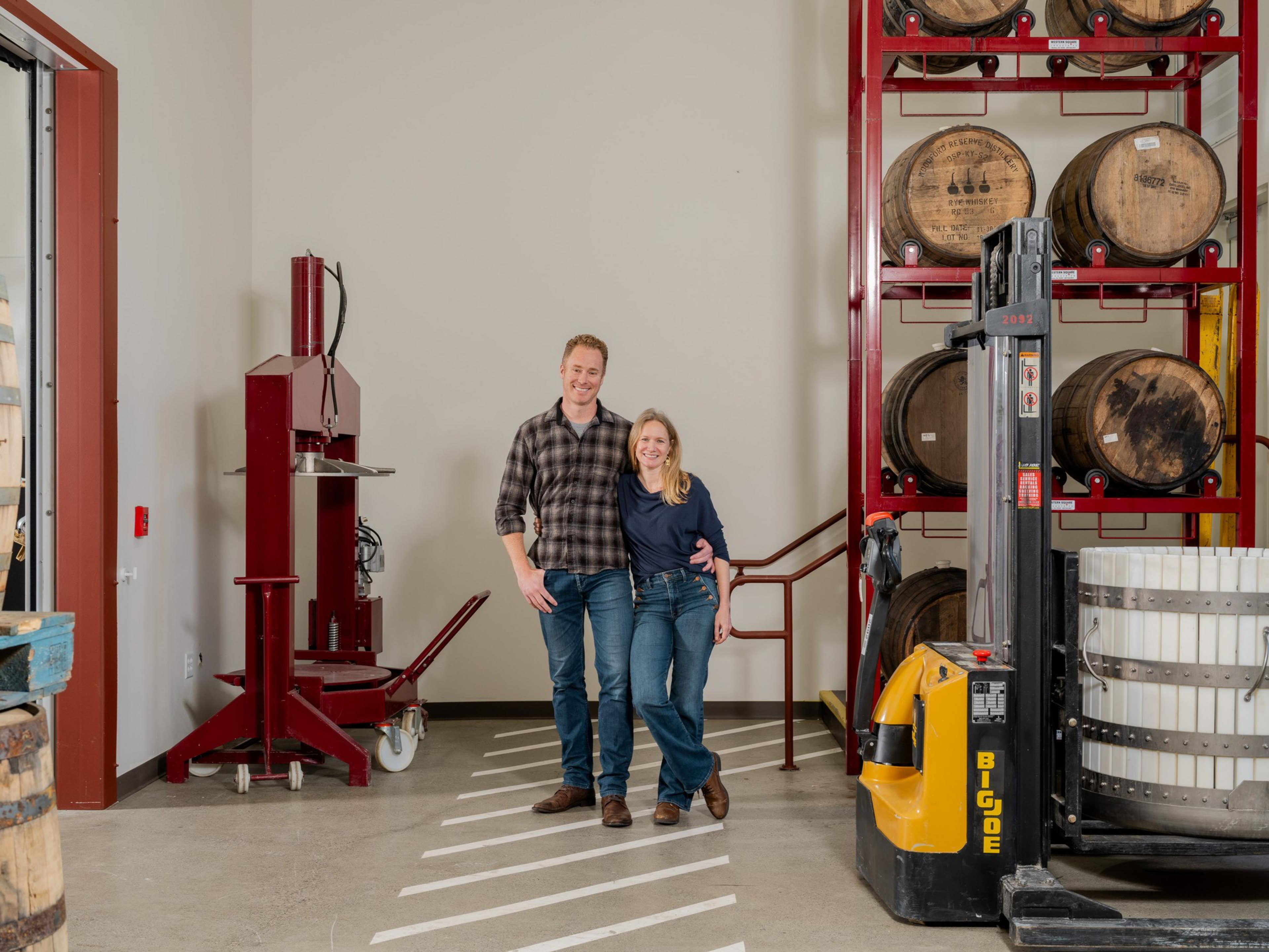 A smiling couple stands in a distillery, surrounded by large wooden barrels on a red rack. They pose near a barrel lifting machine on a concrete floor.