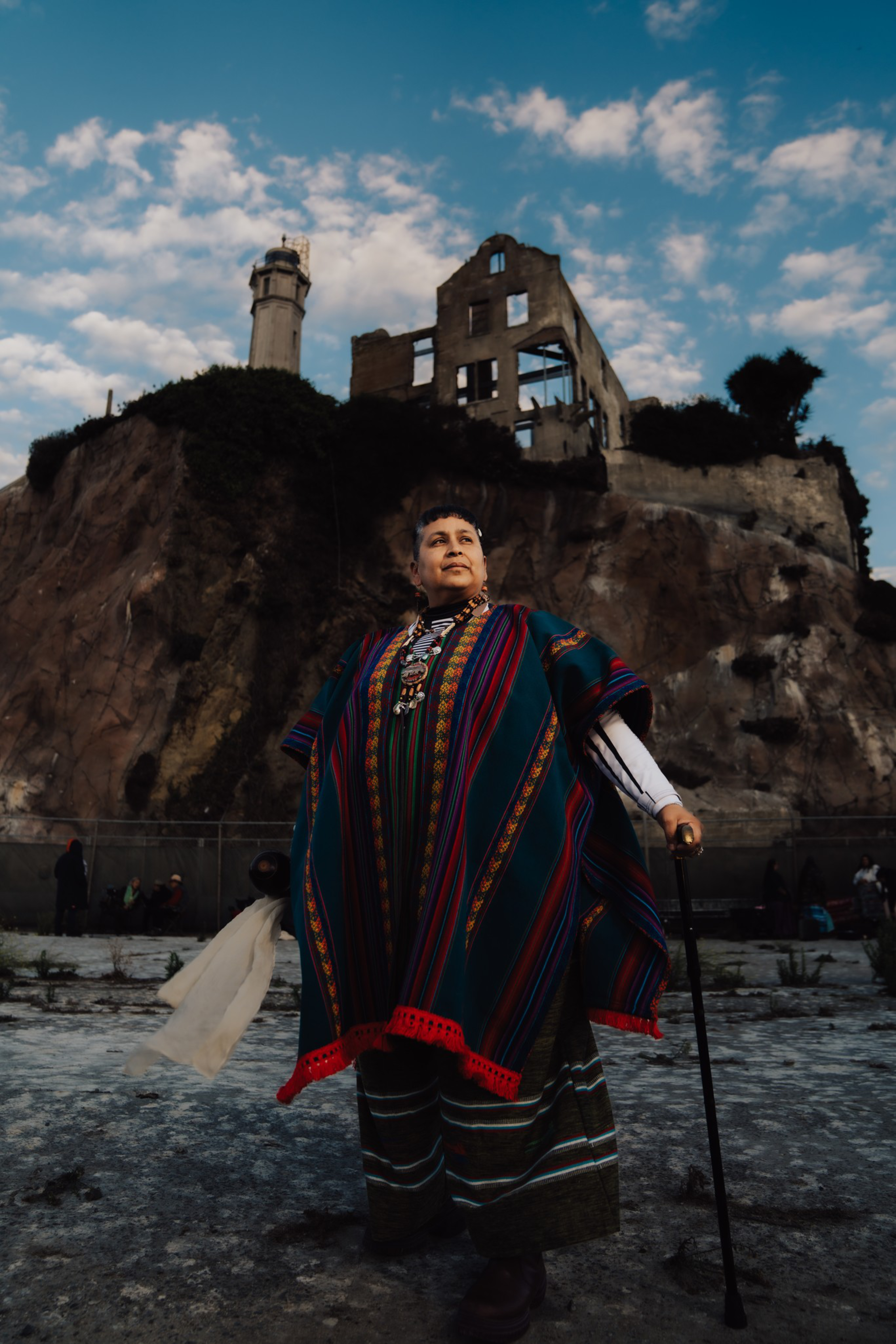 A person wearing colorful traditional clothing stands confidently with a cane in front of a rocky hill topped with a ruined building and lighthouse.