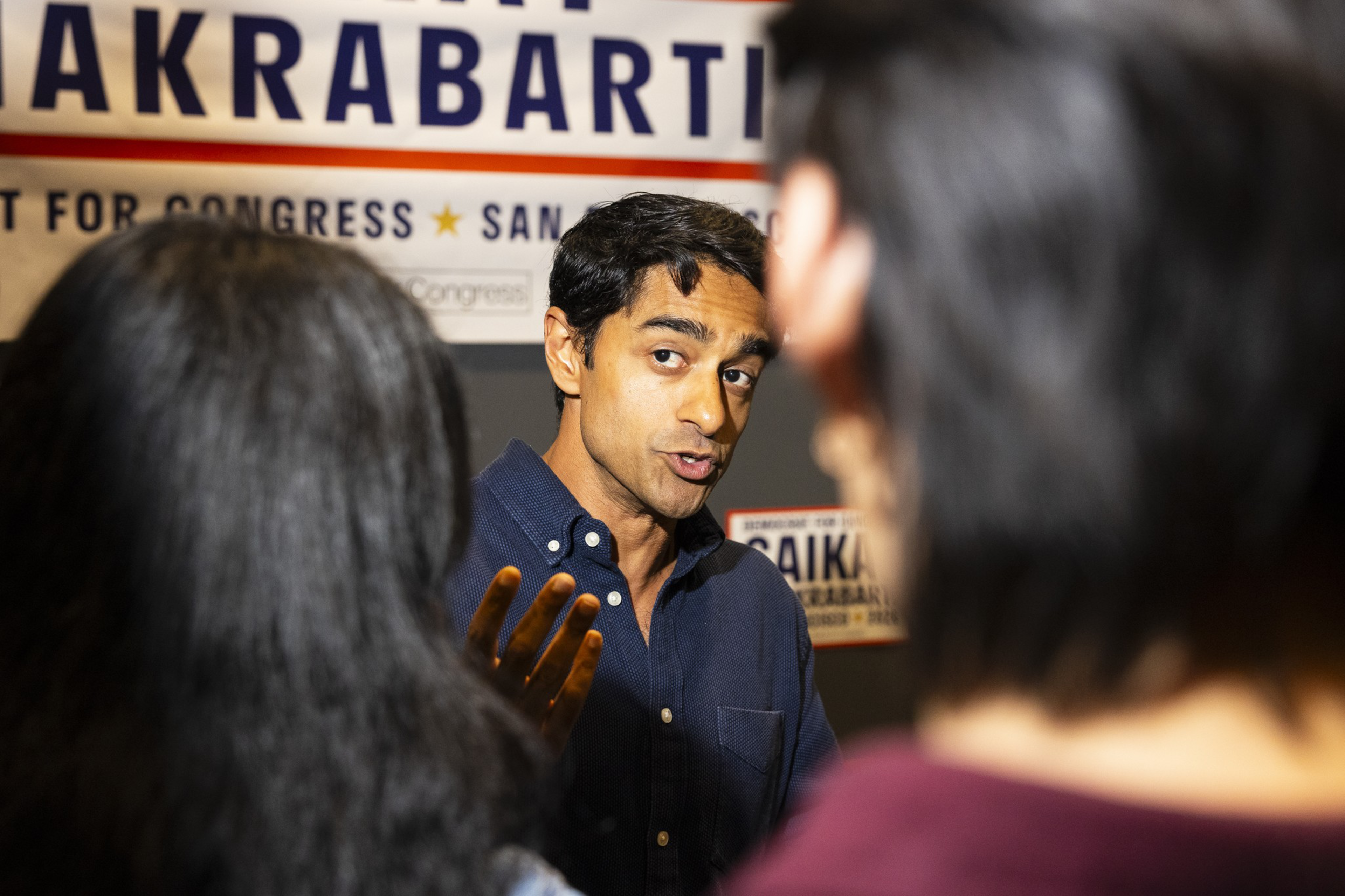 A man in a dark blue shirt speaks animatedly to two people, with campaign signs behind him displaying the name "Saikat Chakrabarti."