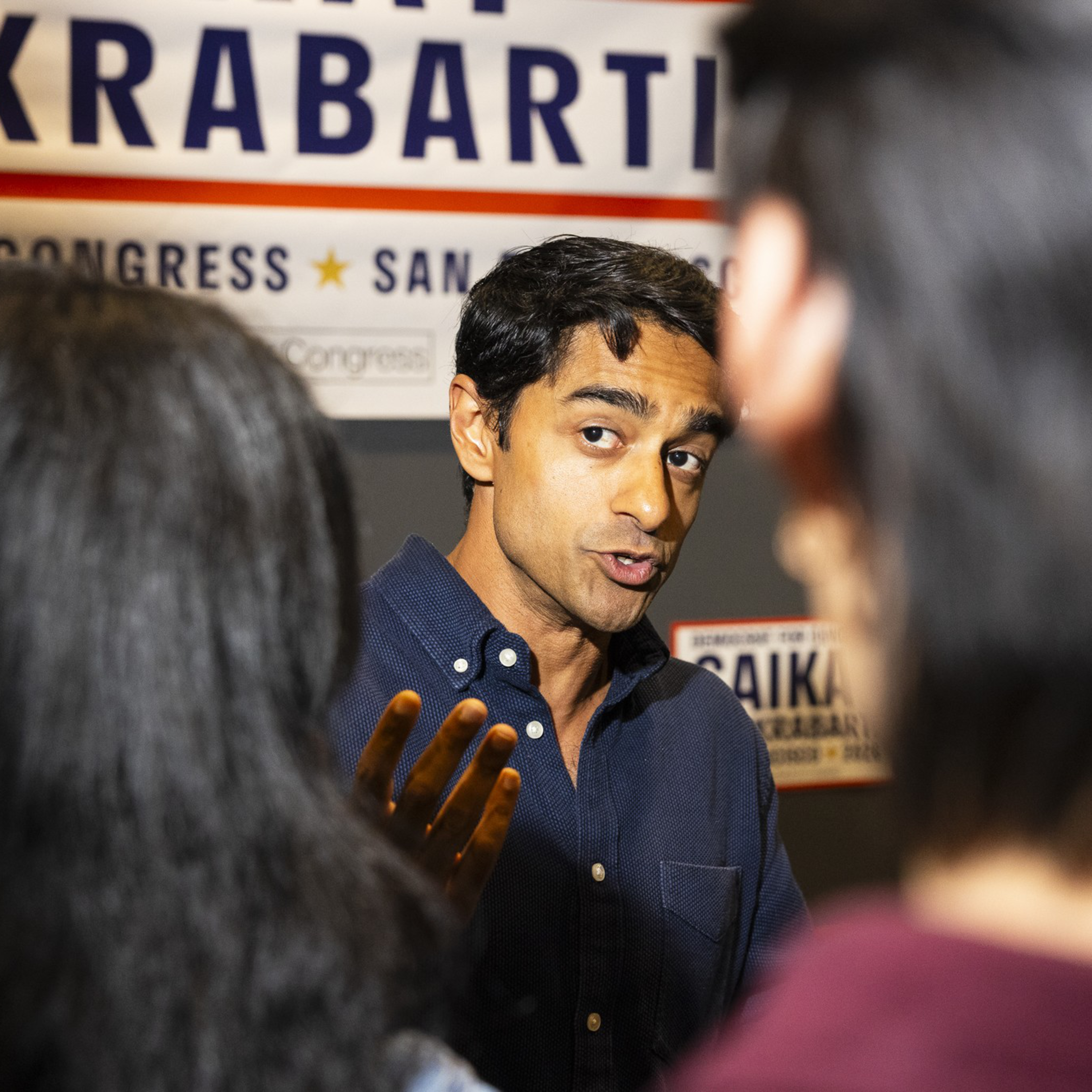 A man in a dark blue shirt speaks animatedly to two people, with campaign signs behind him displaying the name "Saikat Chakrabarti."