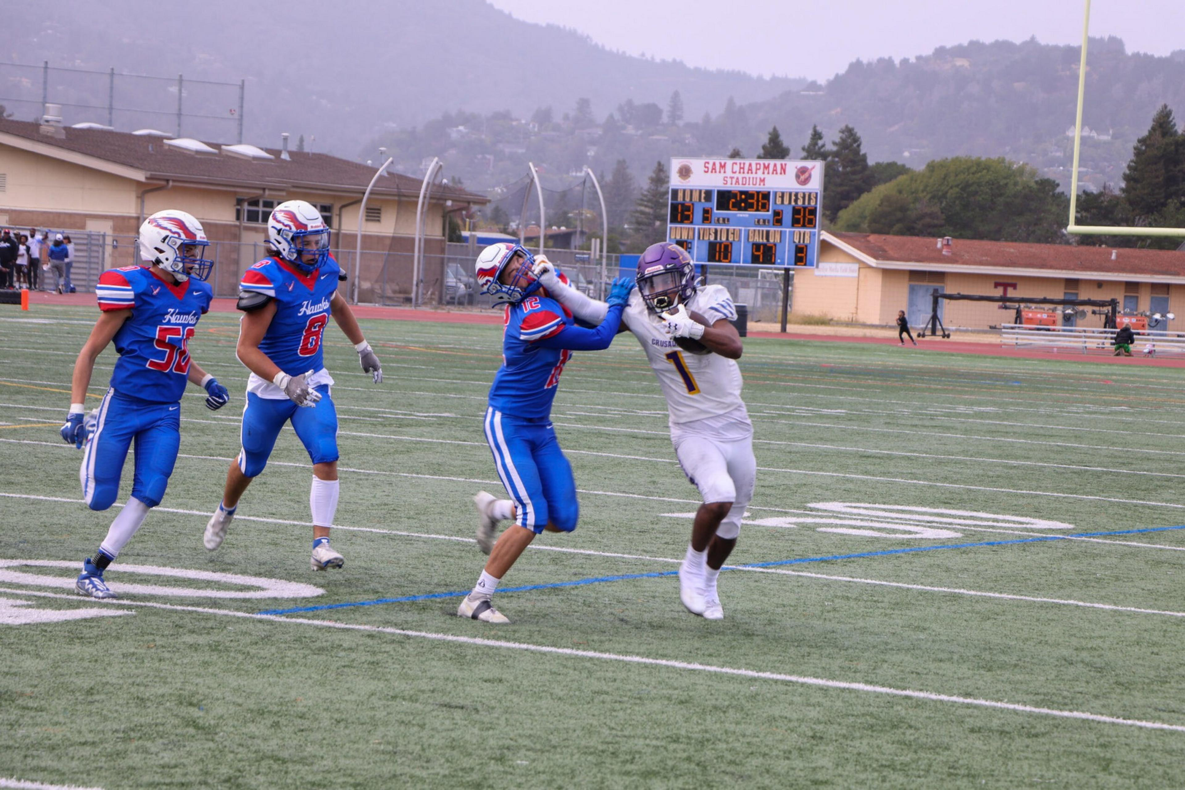 A football player in white with the ball uses his arm to block a blue-uniformed defender's face, while two more defenders run toward them on the field.