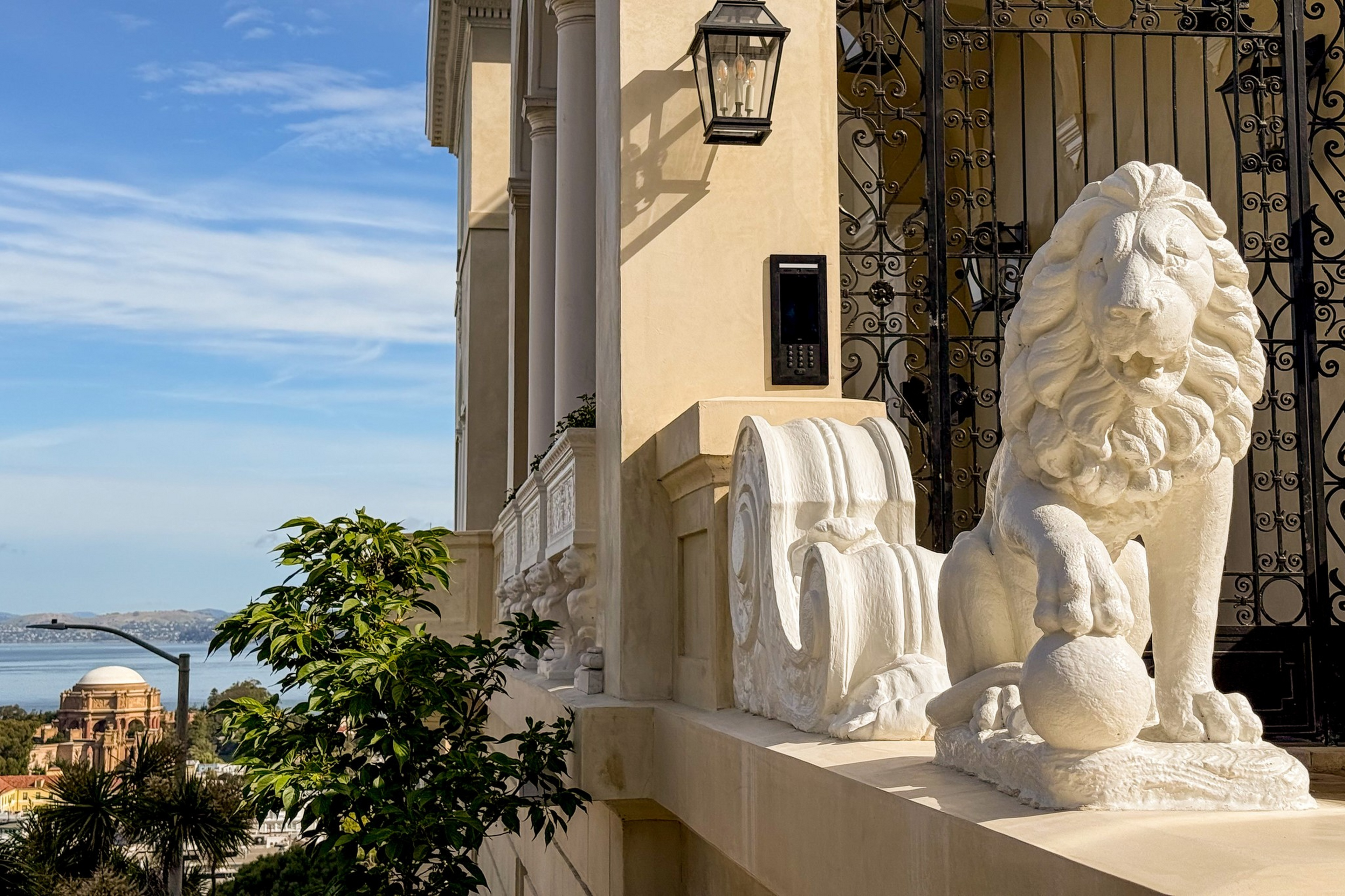 A white stone lion statue with a paw on a ball sits on a building ledge near an ornate black gate, with a scenic water view and distant dome in the background.