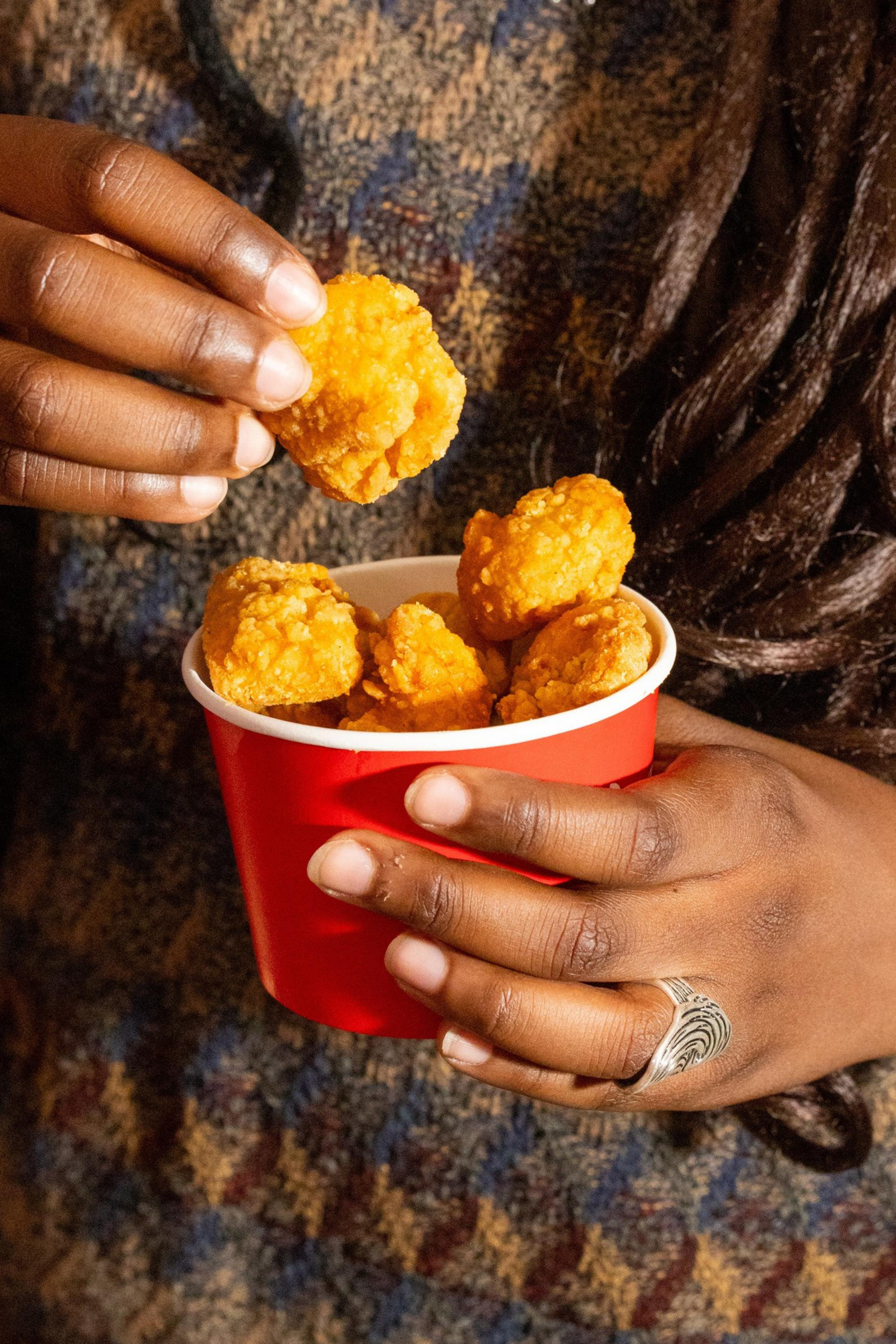 A person wearing a patterned sweater holds a red container filled with golden fried chicken nuggets, picking one up with their fingers.