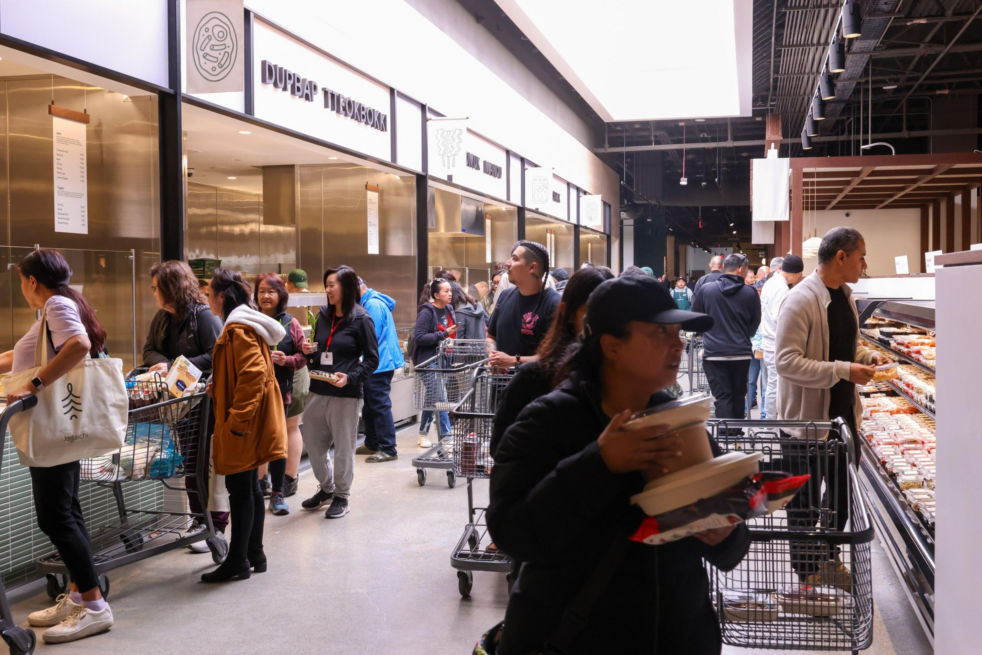 People are shopping in a busy store, each with grocery carts or baskets. They are standing in line at a food counter with signs above.