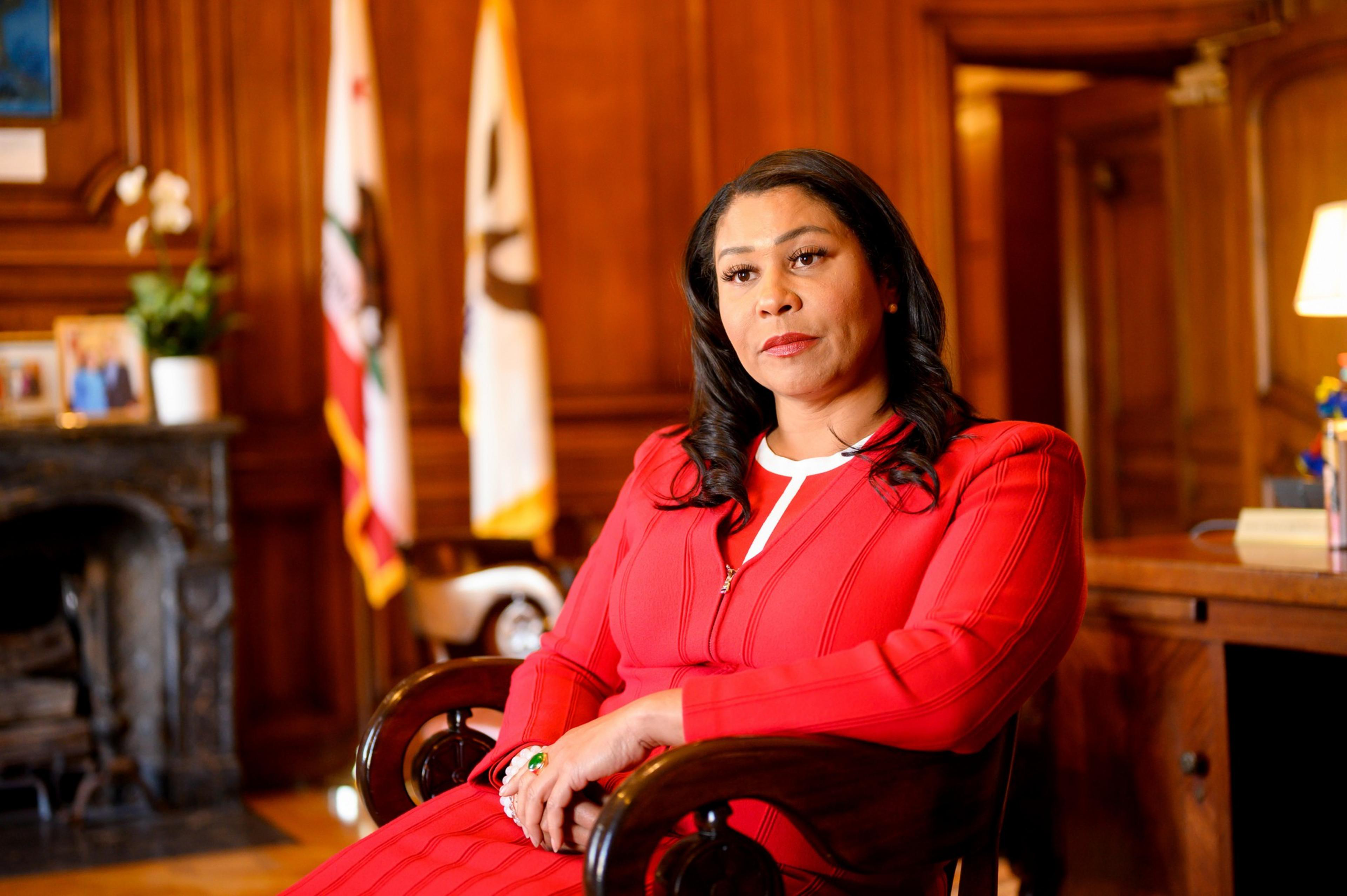 A woman in a red outfit sits solemnly in an ornate wood-paneled room with flags and a fireplace.