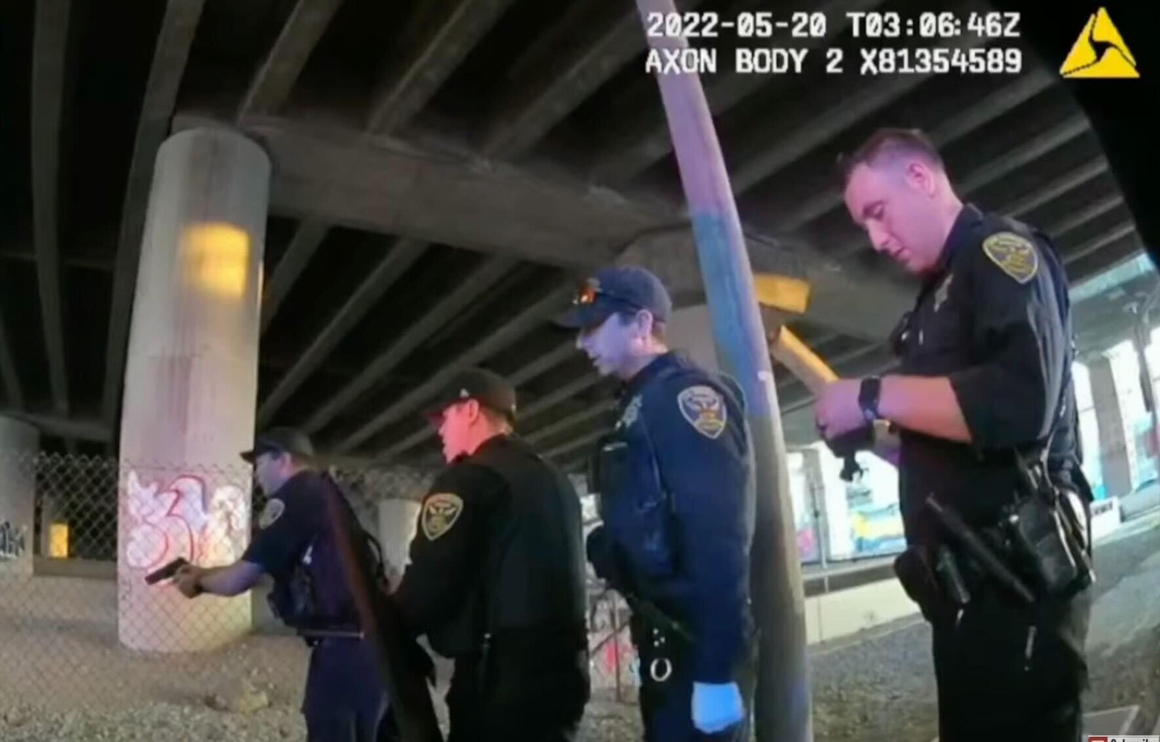 Four police officers stand under a graffiti-covered overpass, two aiming guns forward while the others watch or check equipment.