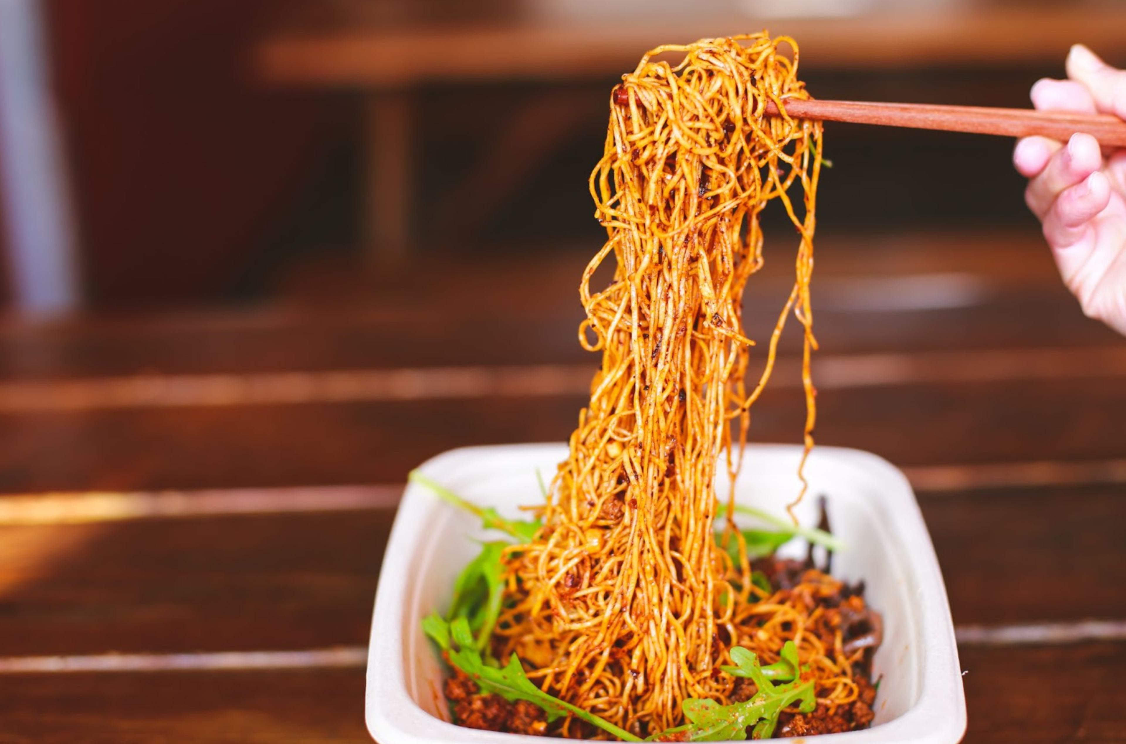 Thin noodles coated in sauce are lifted with chopsticks from a rectangular white container, resting on a wooden table with some green garnish visible.