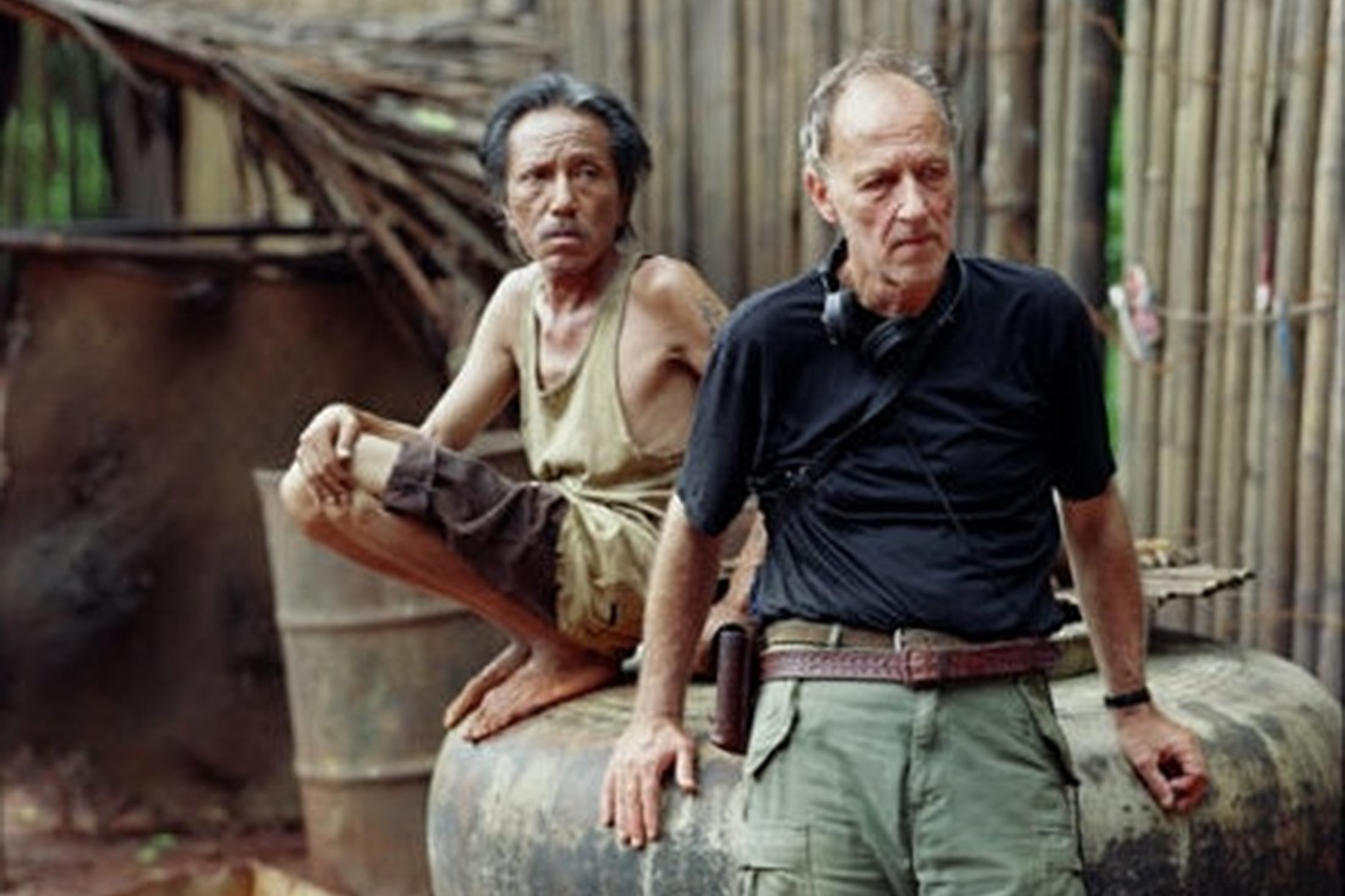 One man in a tank top and shorts squats on a large container while another man in a black shirt and green pants leans on it, both outdoors near wooden structures.